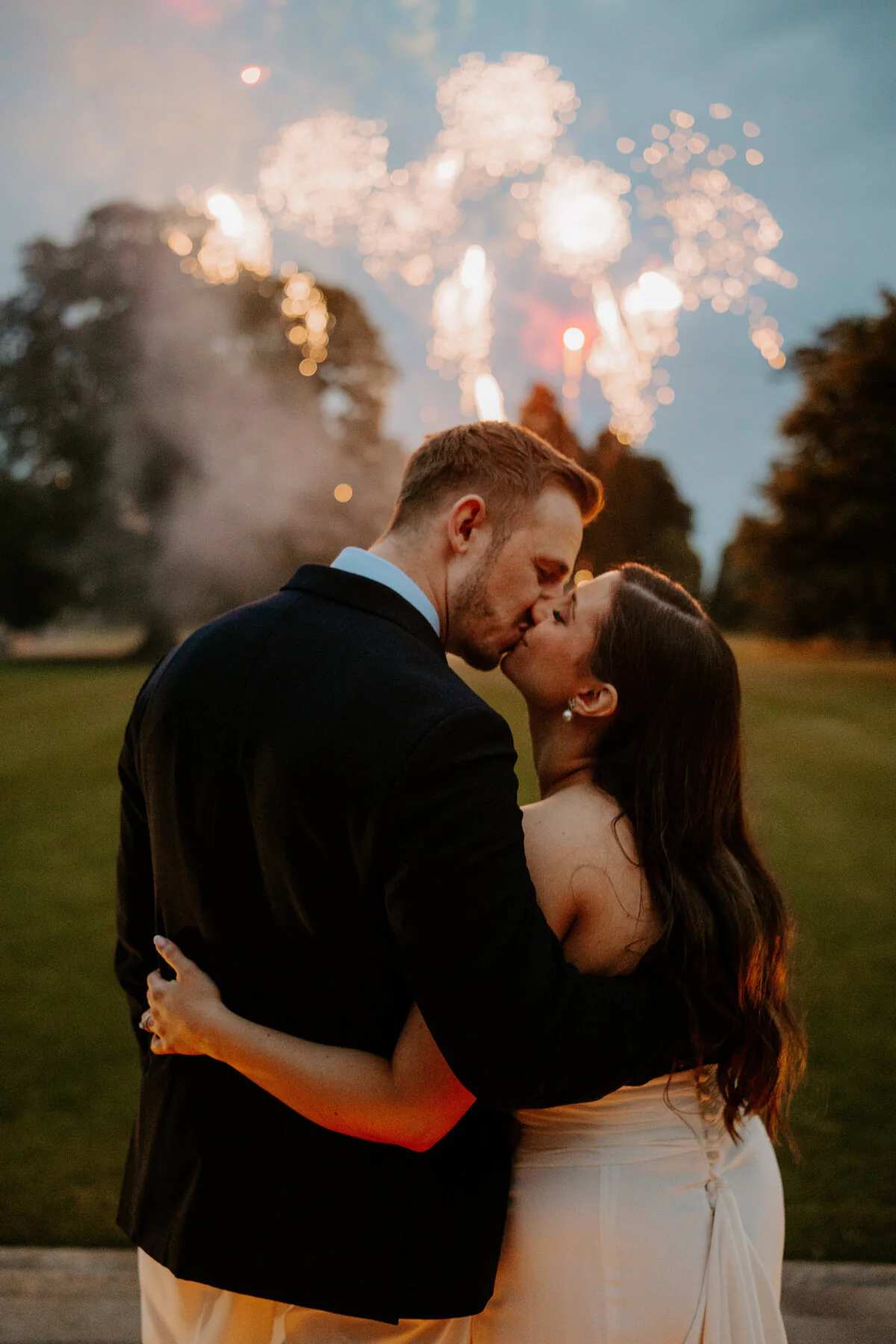 Bride and groom kissing beneath a firework display at night.