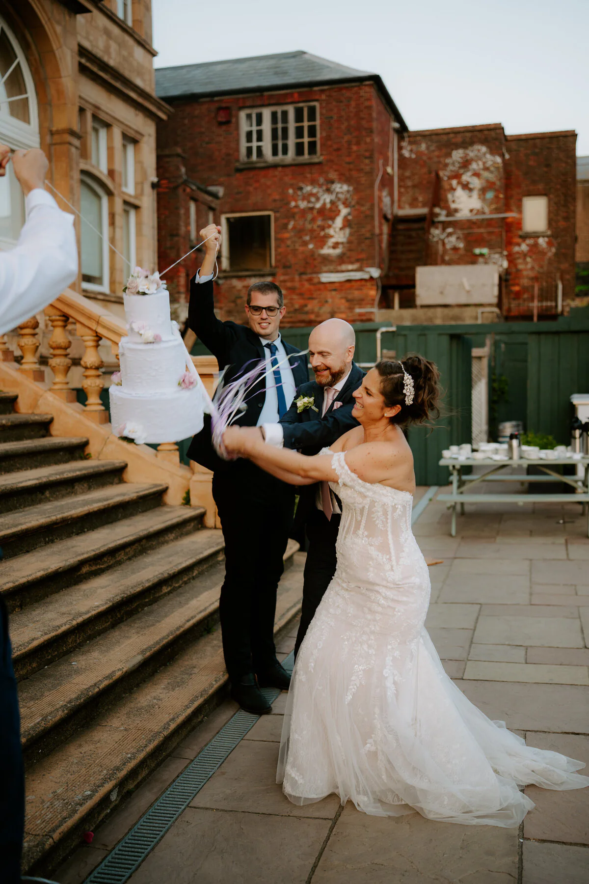 Bride and groom pulling a ribbon to reveal their wedding cake on the venue steps.