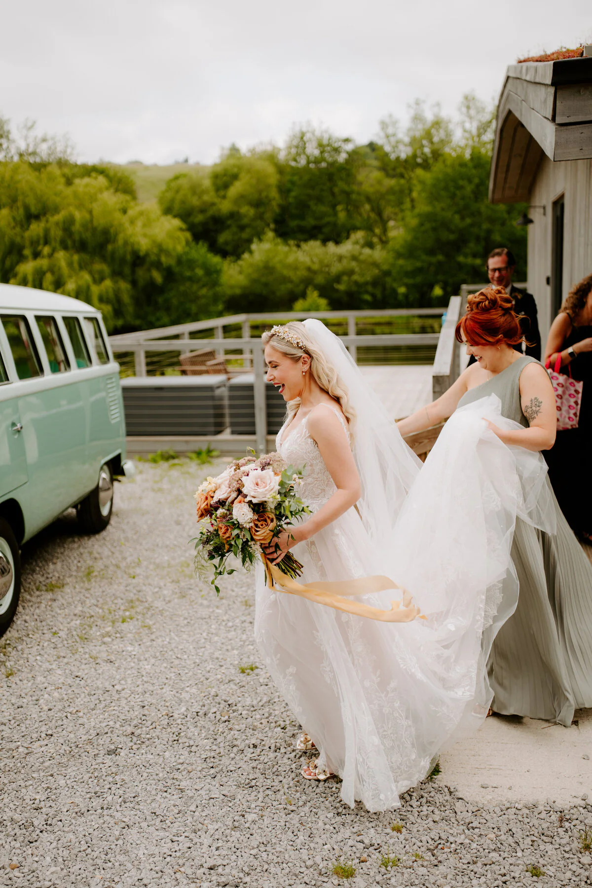 Laughing bride in a lace gown holding an autumn bouquet beside a mint-green VW campervan.