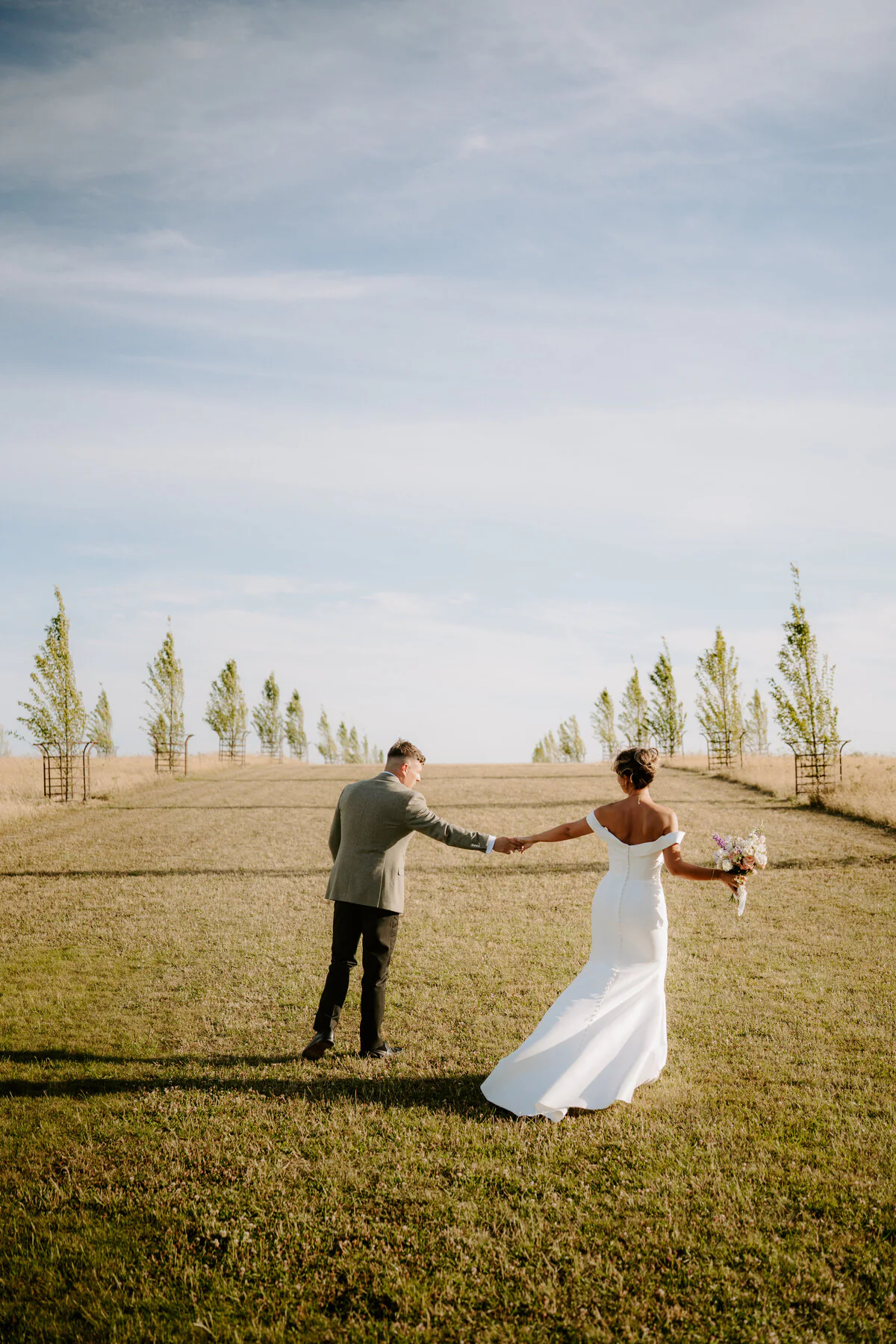 Bride in an off-shoulder gown and groom in grey tweed walking up a grassy hillside path.