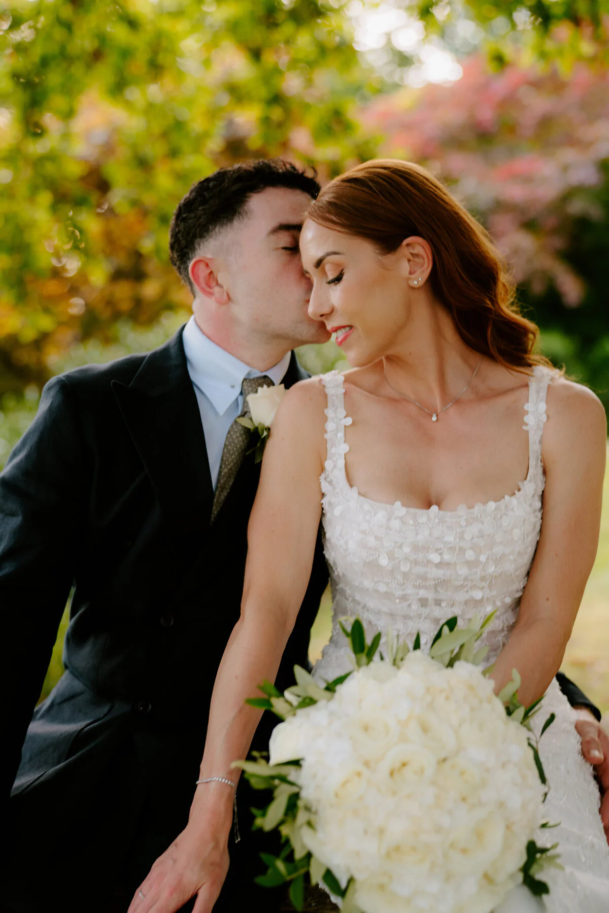 Groom kissing the bride's temple as she holds a white rose bouquet in a floral-applique gown.