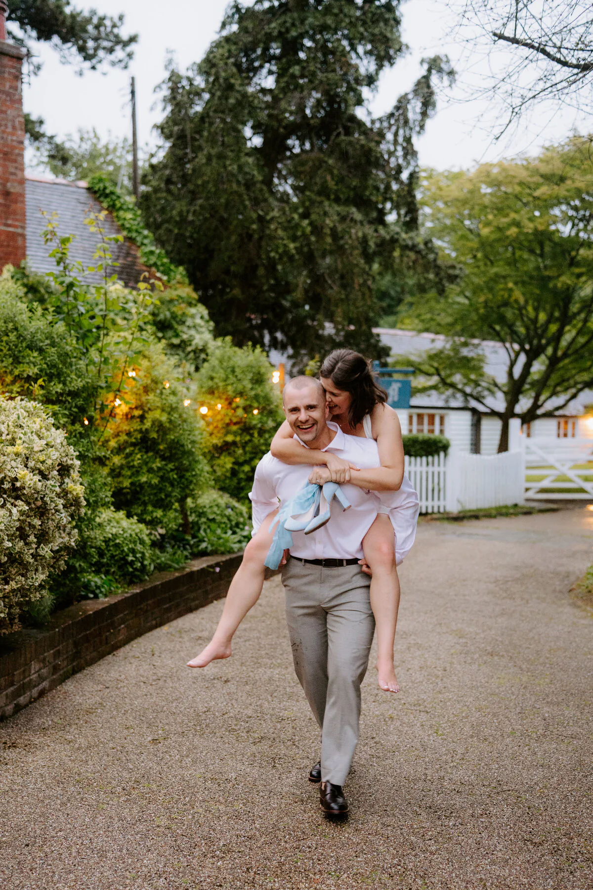 Groom giving the bride a piggyback down a gravel drive past a picket fence.