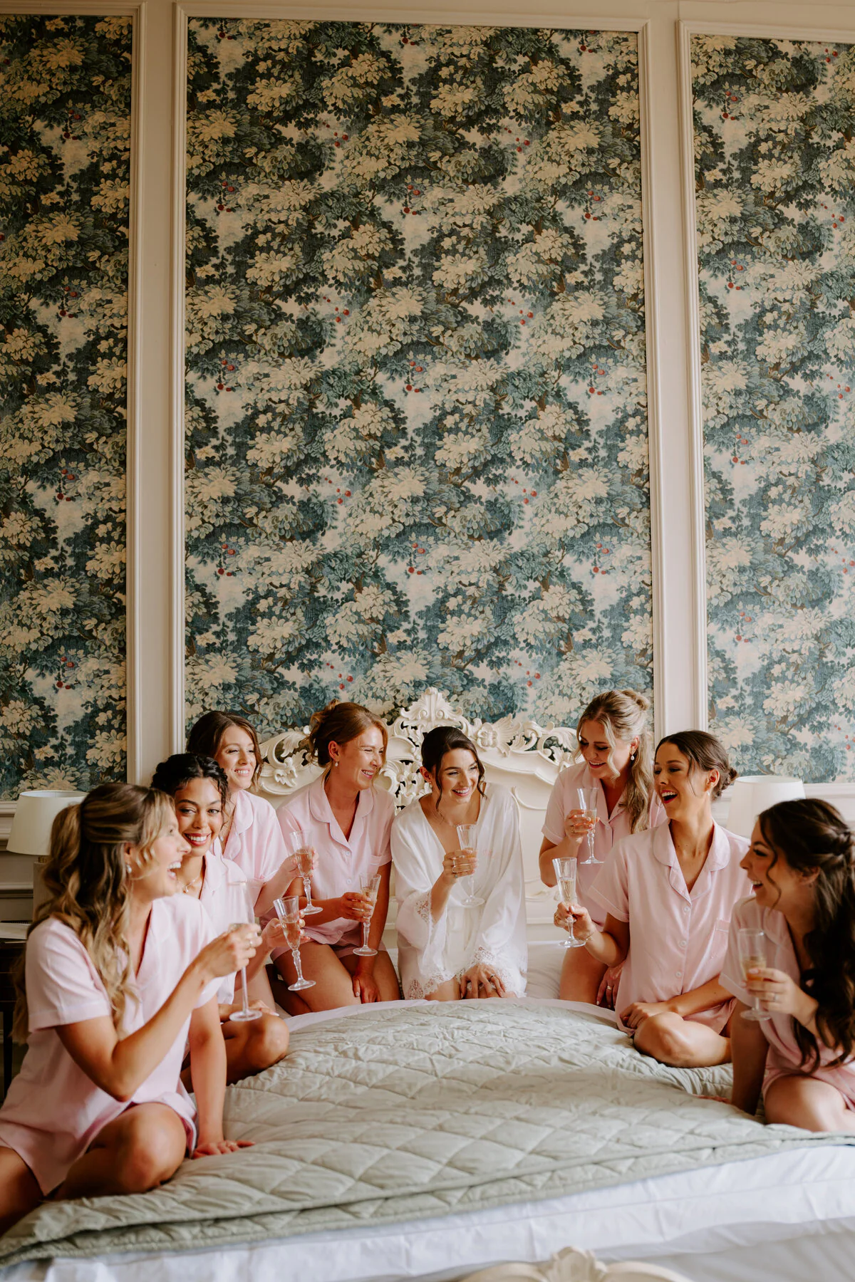 Bride in a white robe with bridesmaids in pink pyjamas on a bed with champagne.