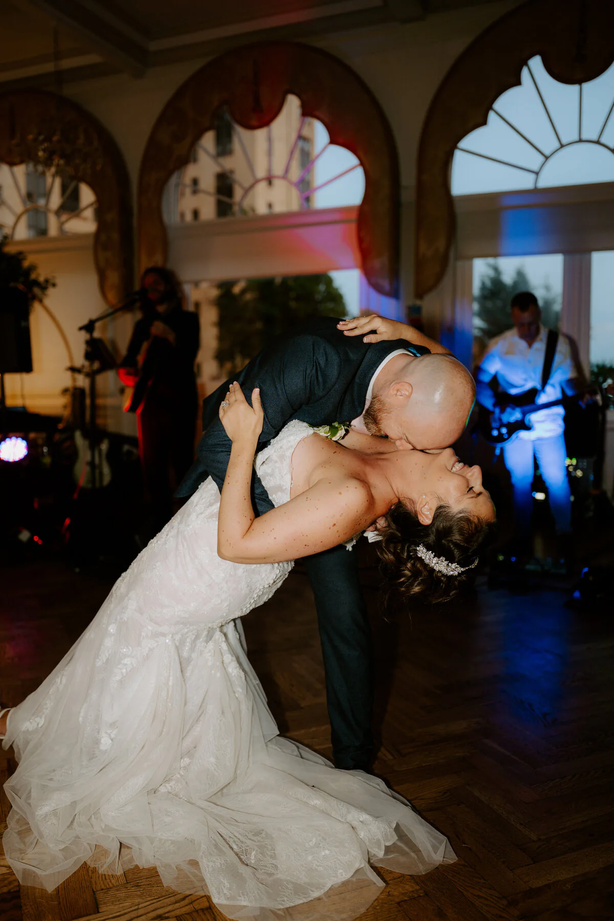 First-dance dip in front of a live band beneath art-deco arched windows.