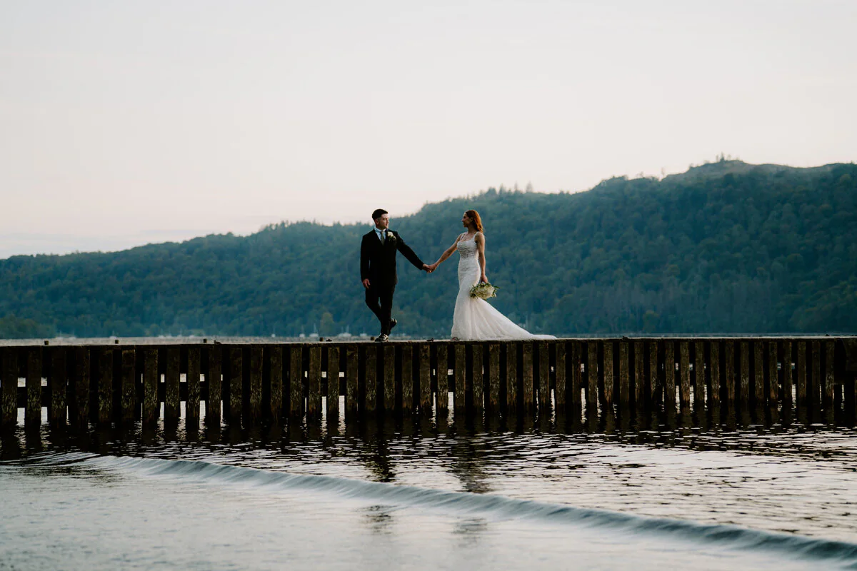 Couple holding hands on a wooden breakwater over a still lake at dusk.
