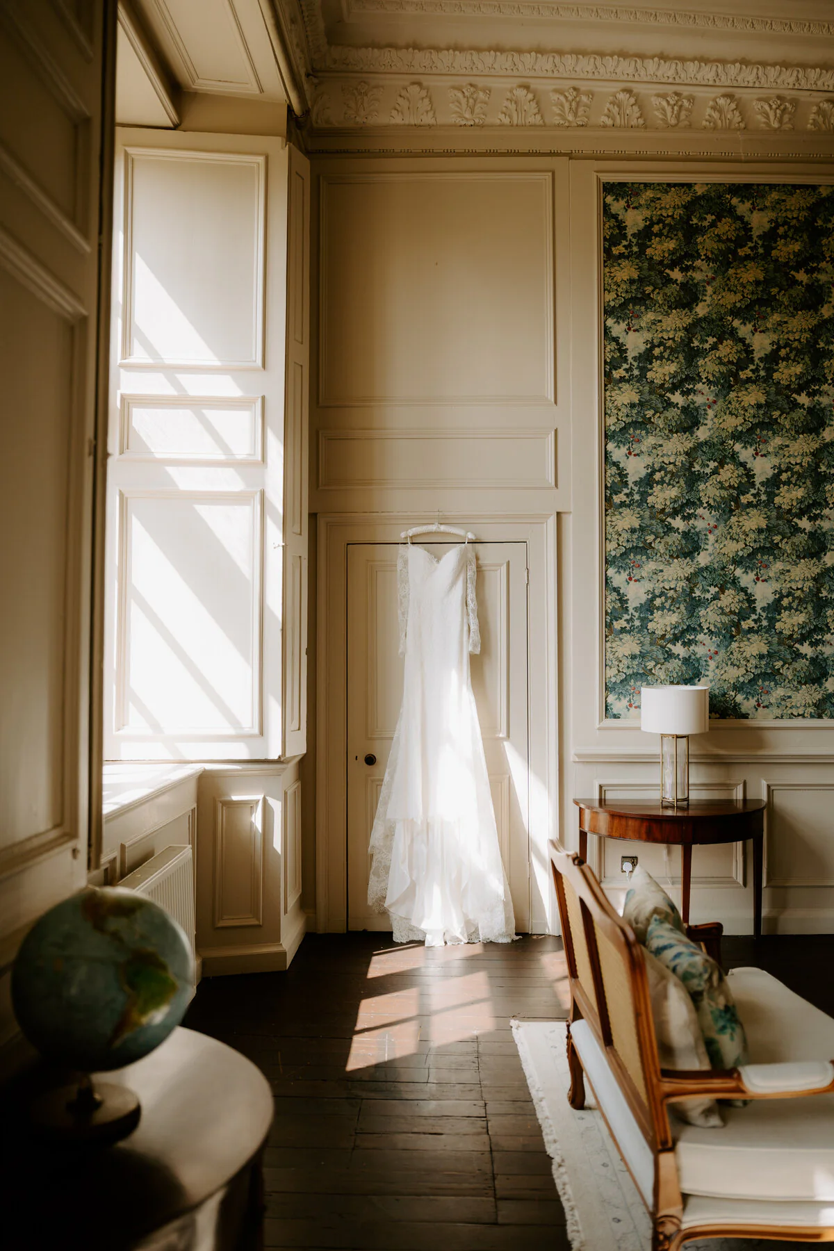 Wedding dress hanging in a sunlit period room with panelled walls and a tapestry.