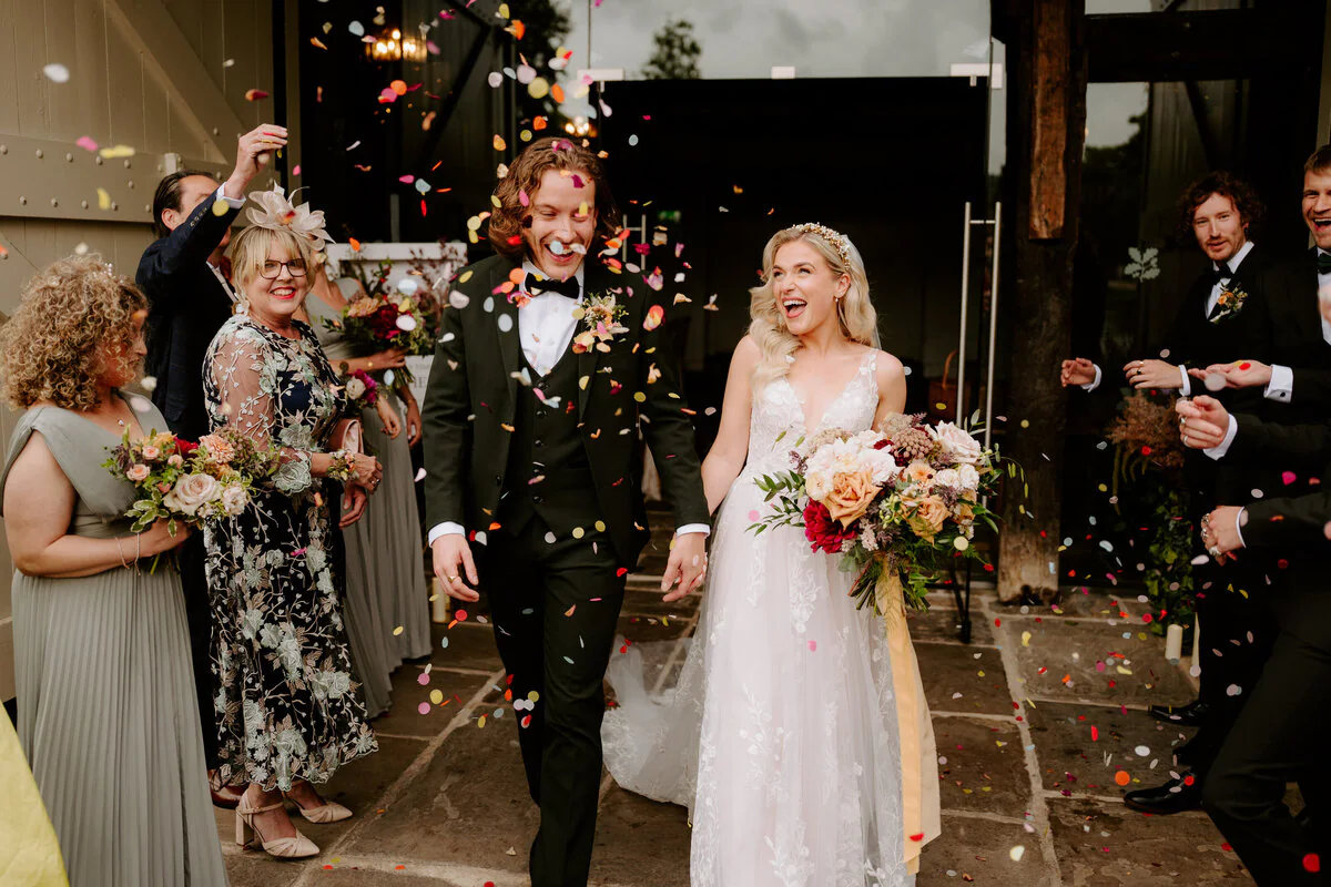 Confetti thrown outside a barn entrance over a lace gown and dark-green three-piece suit.