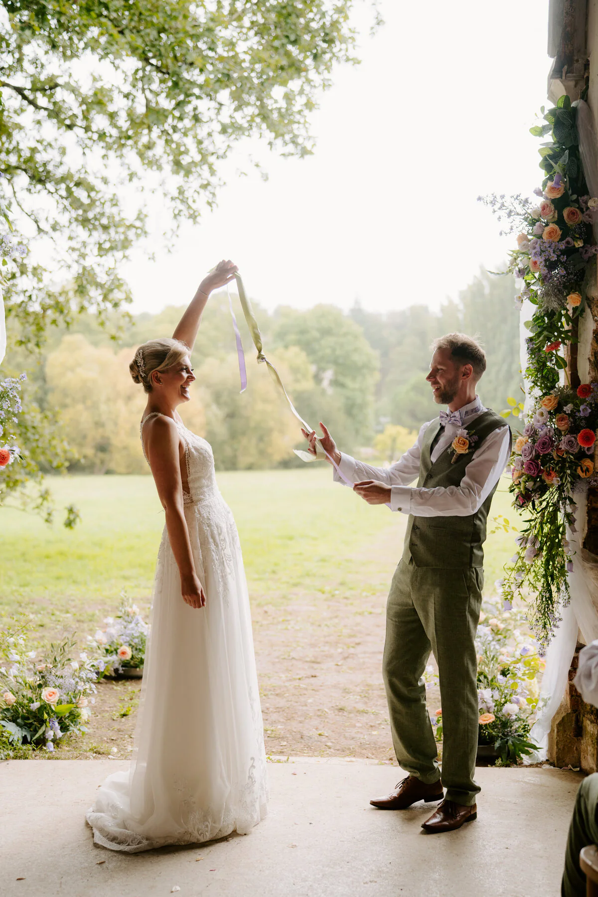 Couple playing a handfasting ribbon game in a barn doorway beneath a floral arch.