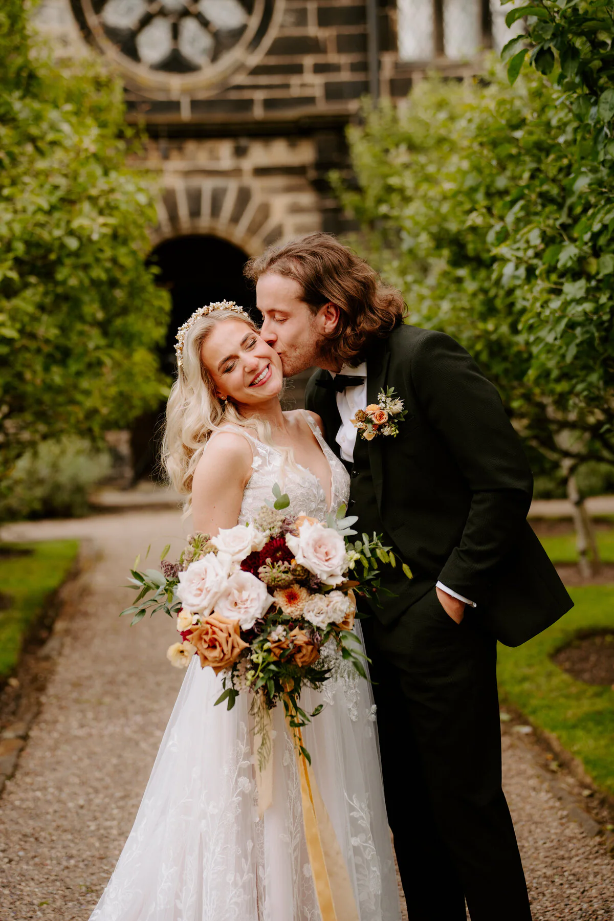 Groom kissing the bride's cheek in a walled garden at a stone manor house.