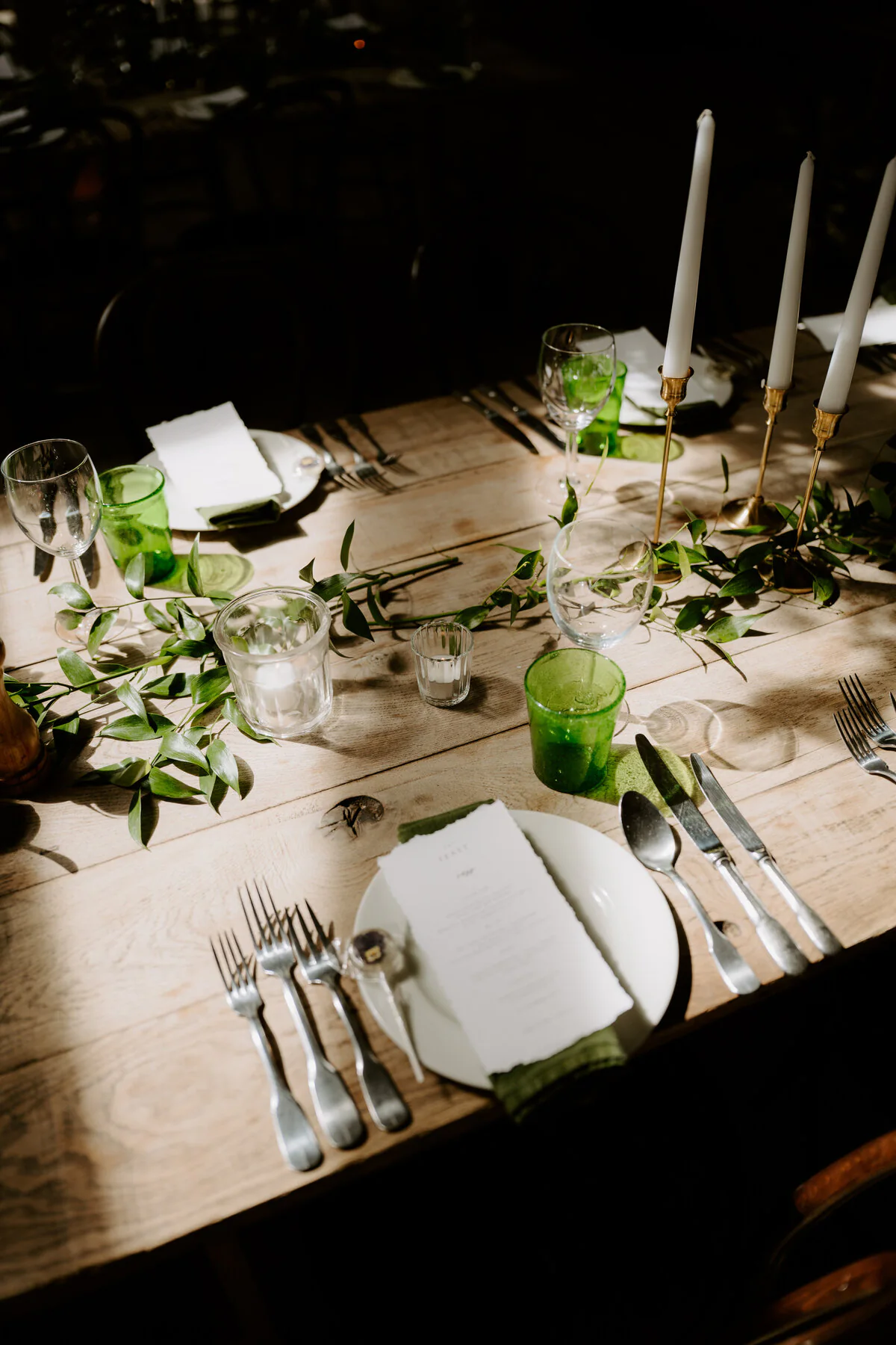 Rustic wooden wedding breakfast table with a greenery runner, candles and green glassware.