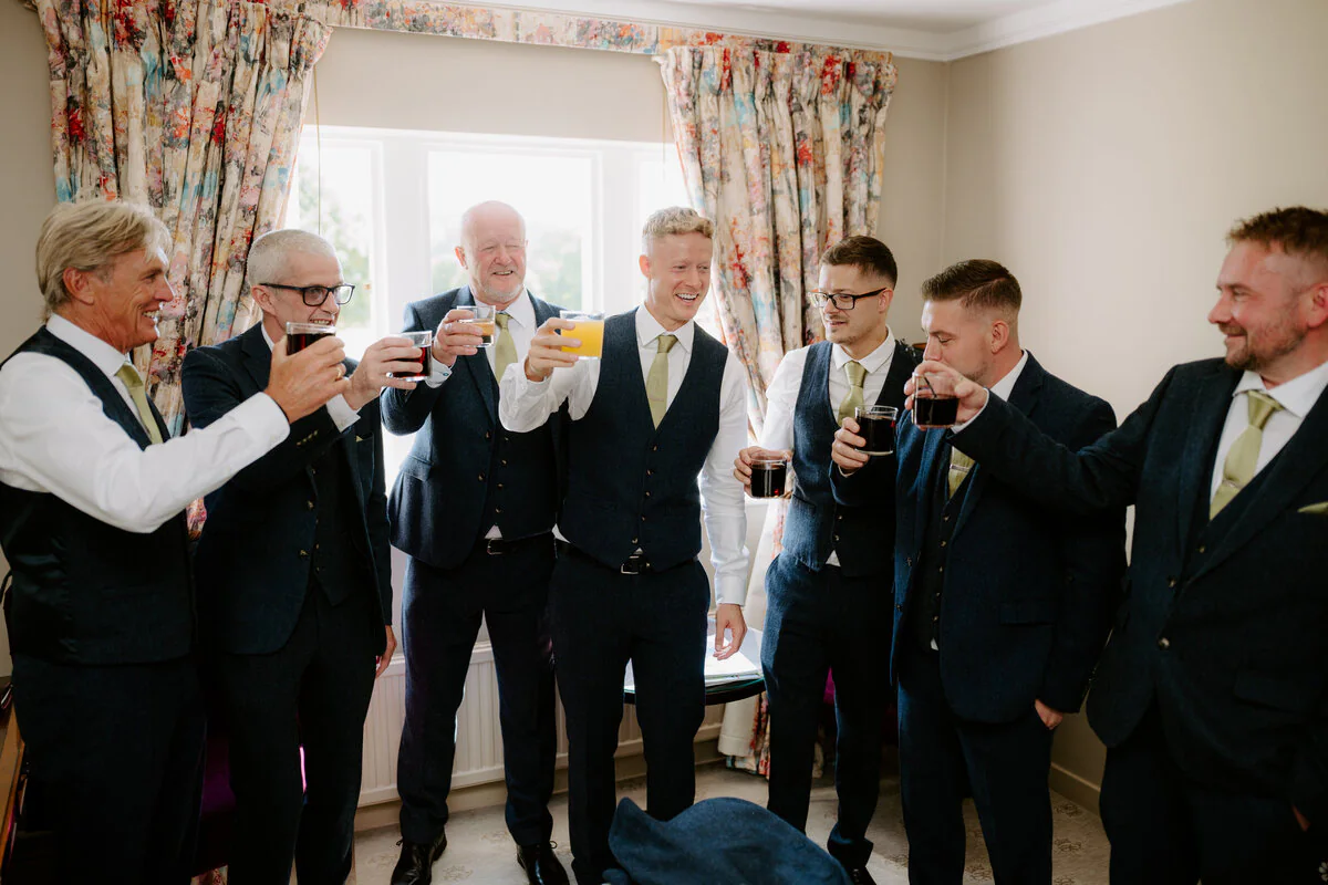 Groomsmen in navy suits toasting with drinks in a cottage drawing room.