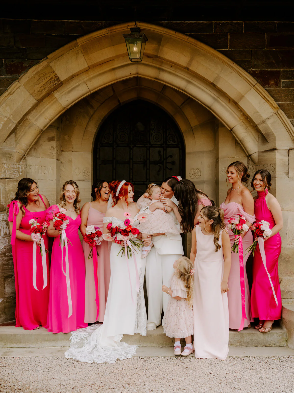 Two brides with their bridal party in pink dresses outside a stone church archway.