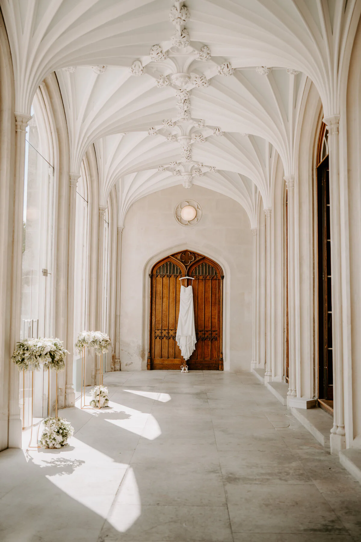 Wedding dress hanging on a gothic wooden door in a white stone cloister corridor.