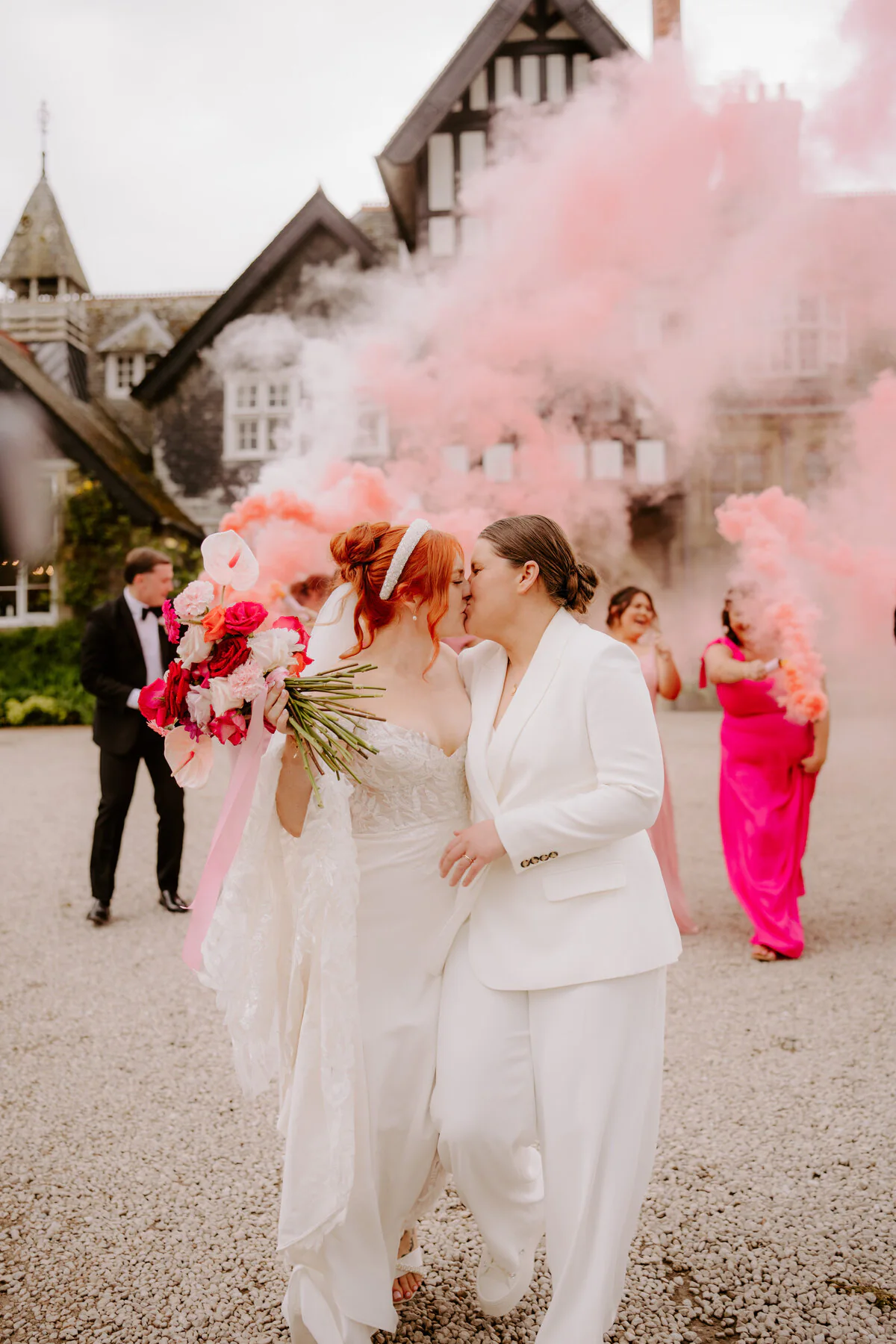 Two brides kissing behind pink smoke bombs outside a Tudor manor with a red bouquet.