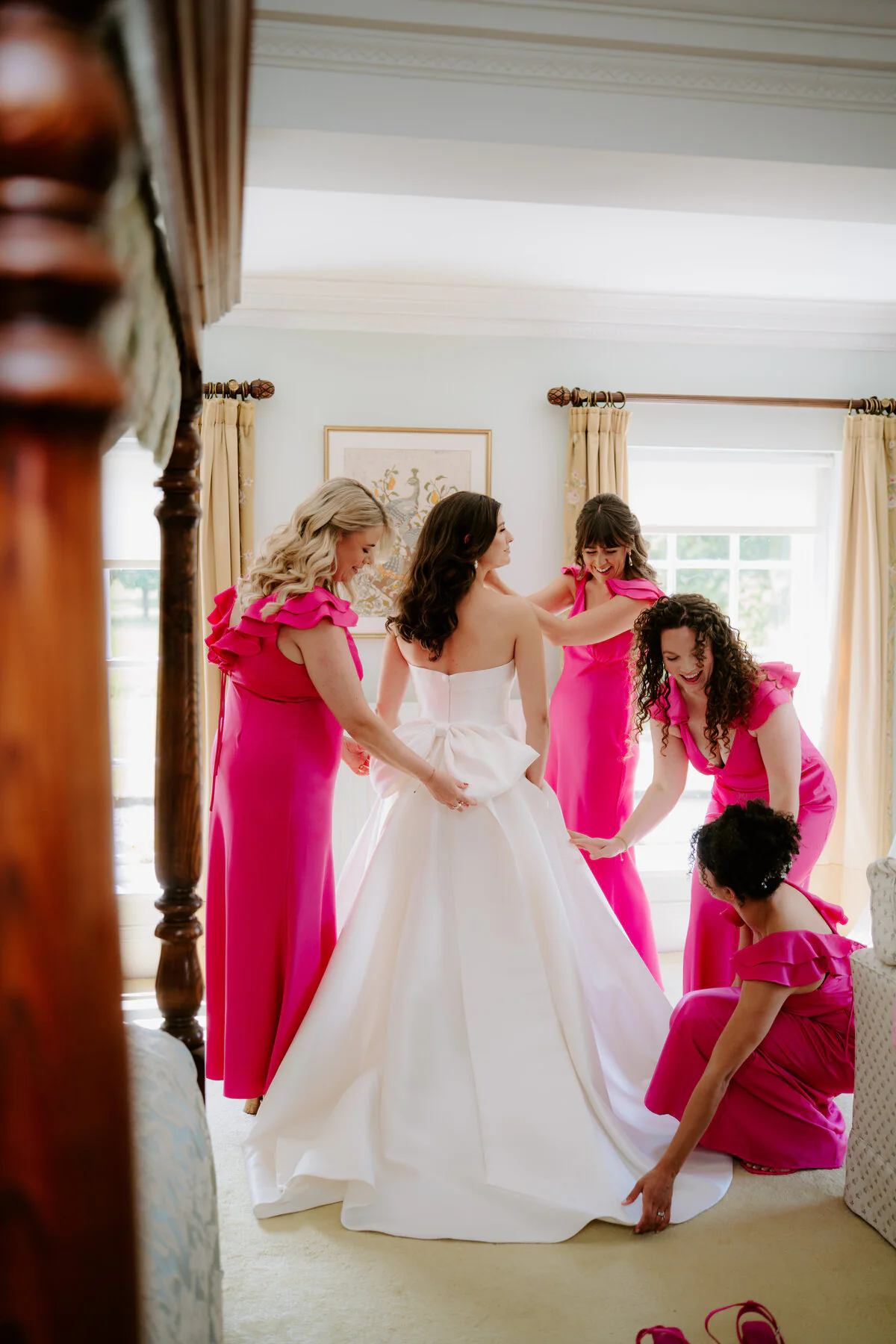 Bridesmaids in hot-pink dresses helping the bride into her bow-back ballgown.