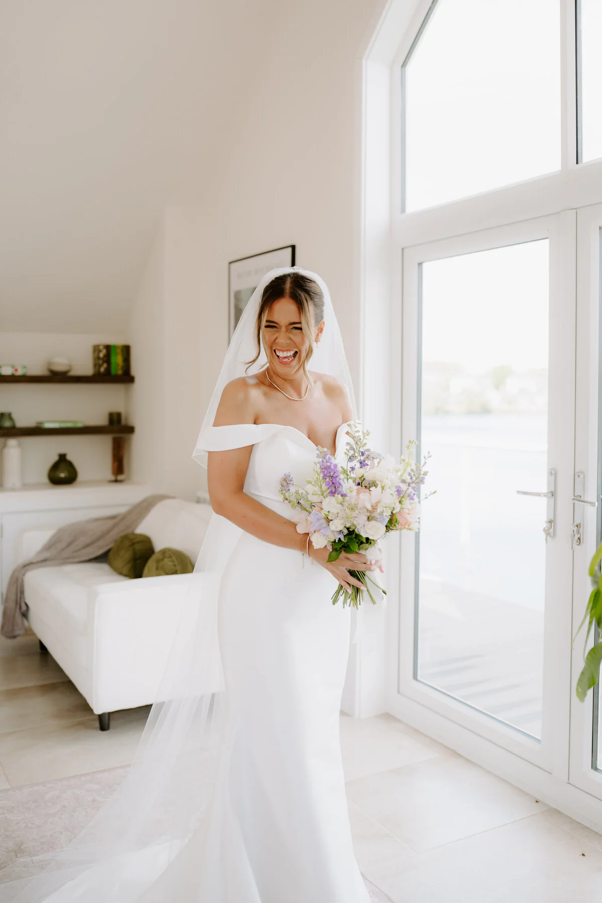 Laughing bride in an off-shoulder gown with a pastel bouquet in a bright lakeside room.