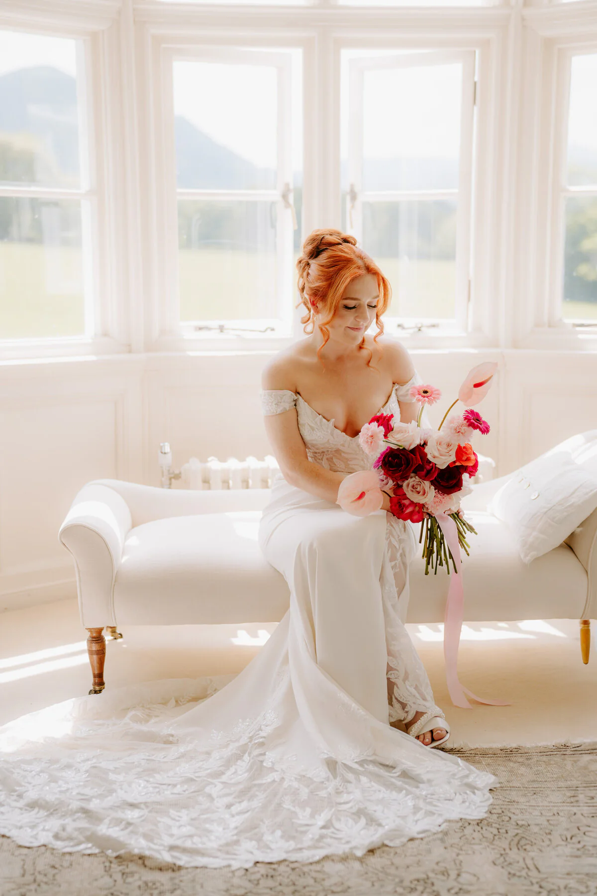 Red-haired bride on a chaise longue with red and pink flowers by a bay window.