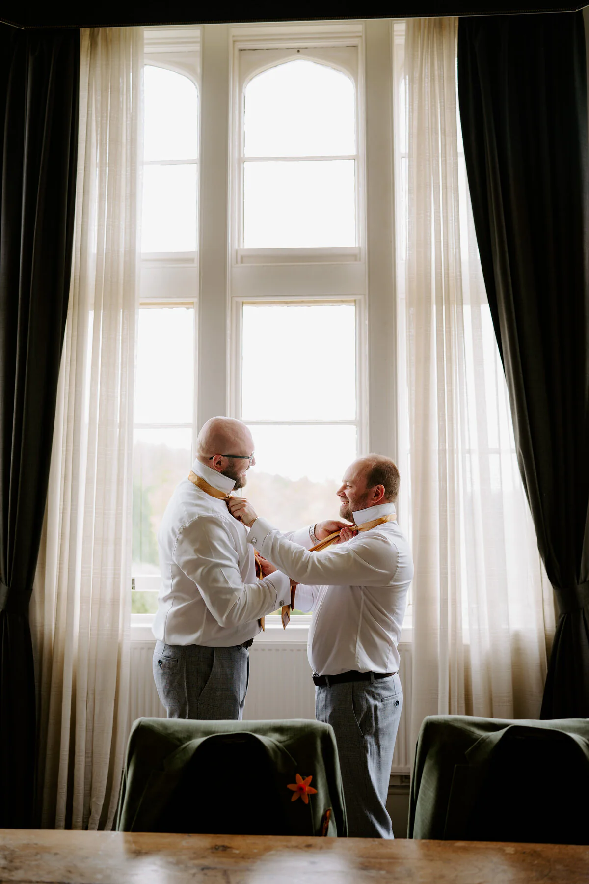 Best man and groom in gold ties getting ready beside a tall window.
