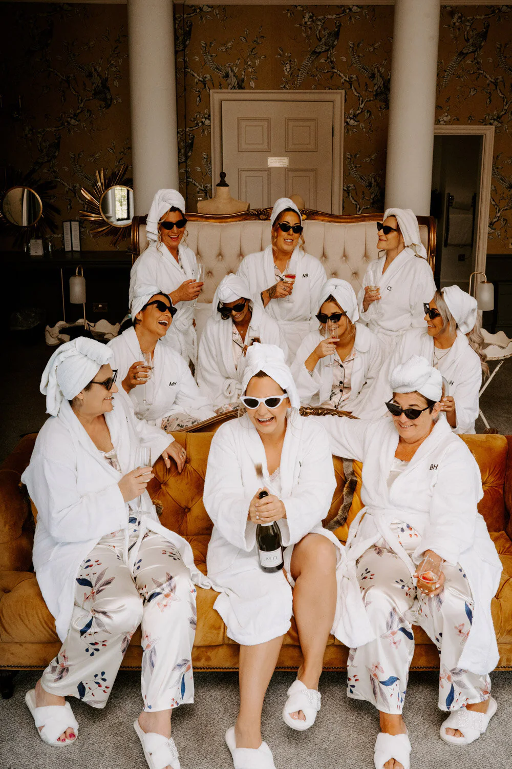 Bridal party in robes with towels on their heads toasting champagne on a velvet sofa.