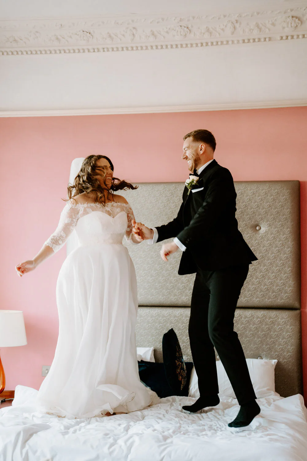 Bride and groom jumping on a bed laughing in a pink bedroom.