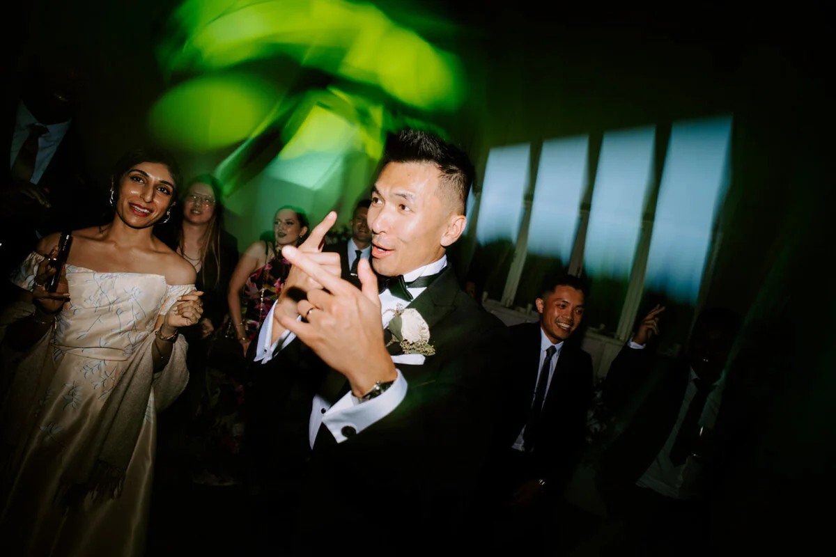 Groom dancing and pointing at the camera under green lights with the bride behind him.