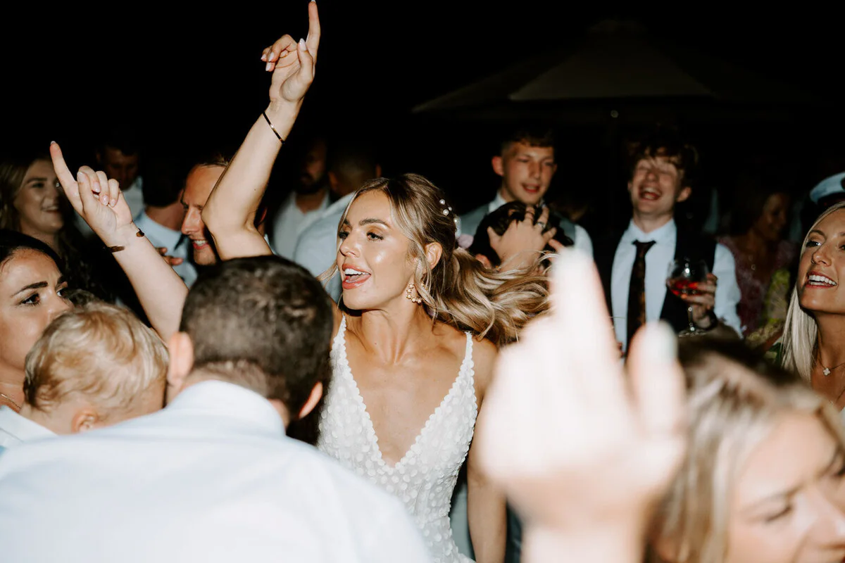 Bride dancing with her arms raised among guests on the dance floor at night.