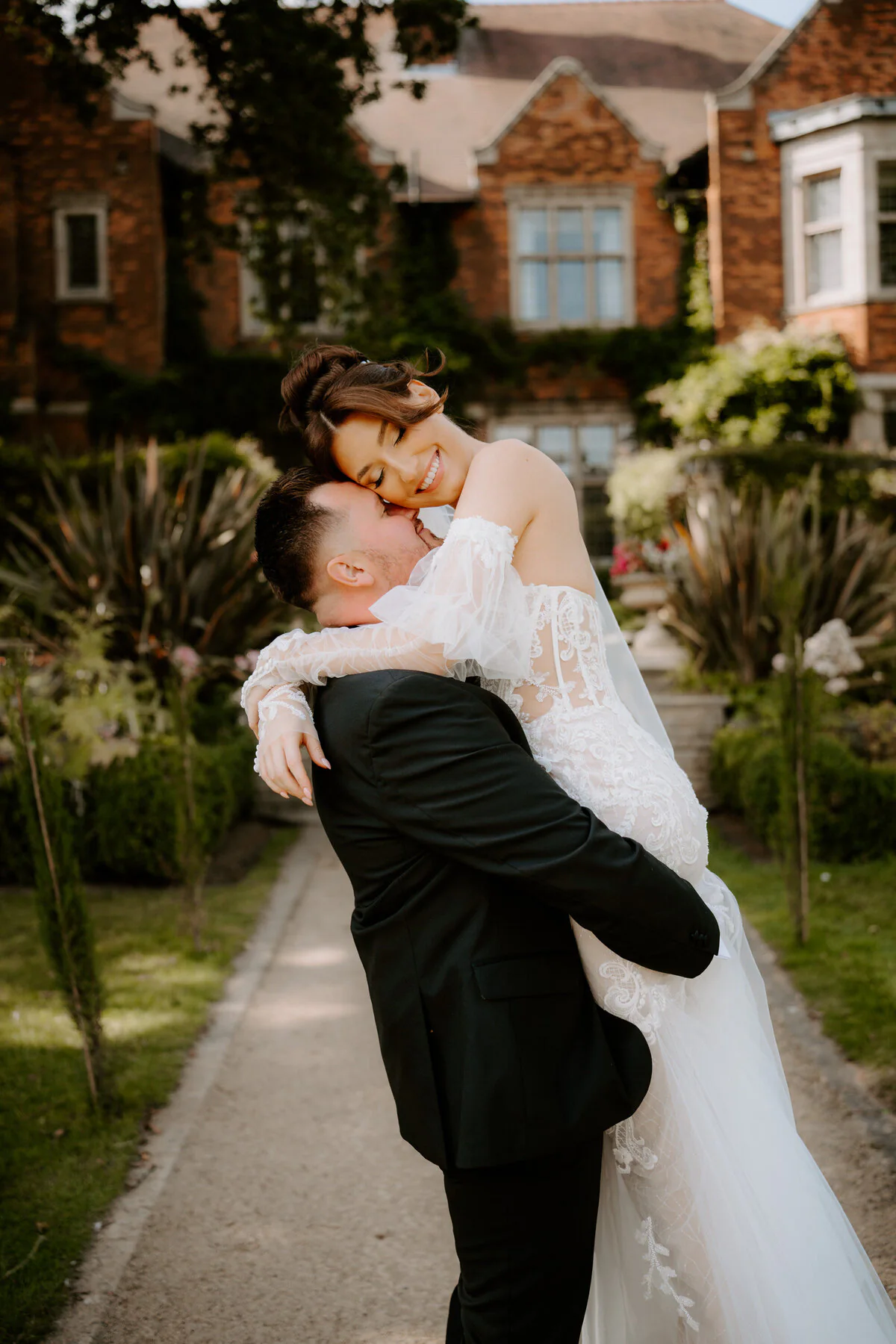 Groom lifting his laughing bride in an off-shoulder lace gown on a manor garden path.