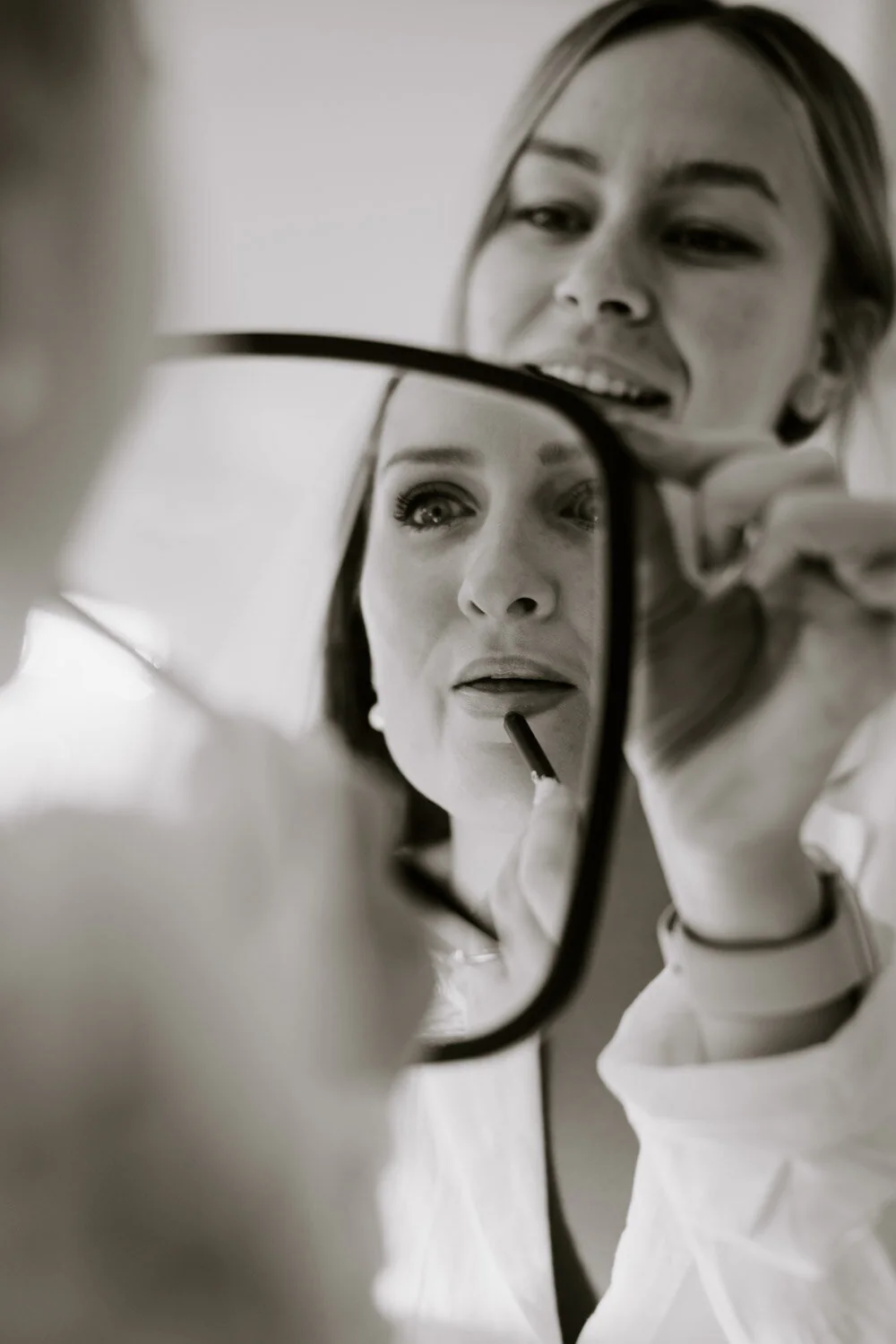 Black and white photograph of a bride applying lipstick in a hand mirror.