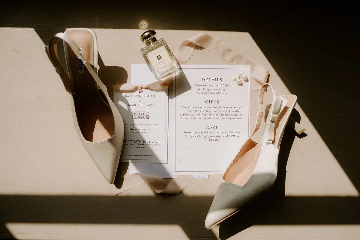 Flat lay of white satin heels, perfume and Katherine and Marcus's wedding stationery in sunlight.