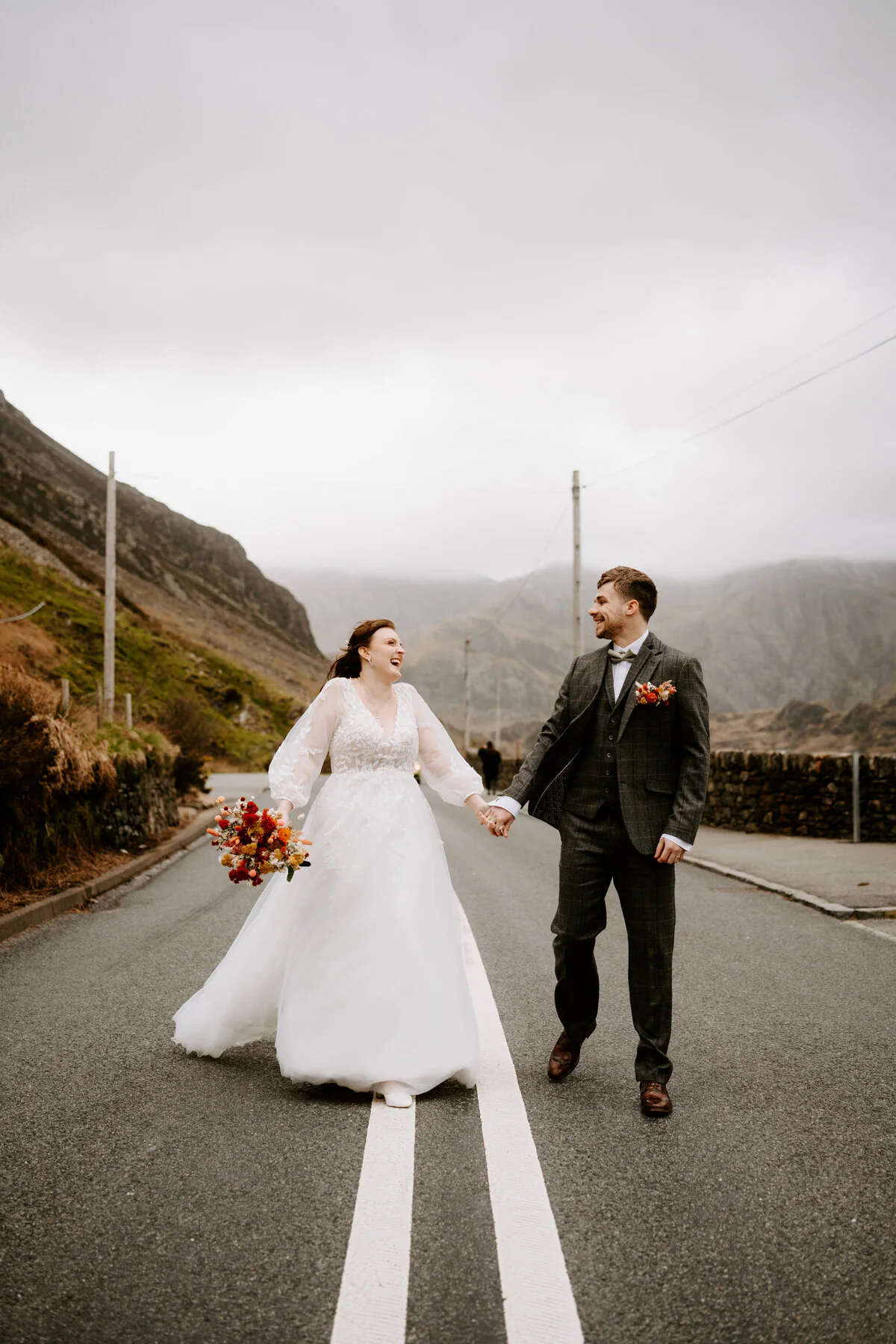 Couple laughing as they walk the centre line of an empty mountain road, with an orange bouquet and tweed three-piece suit.
