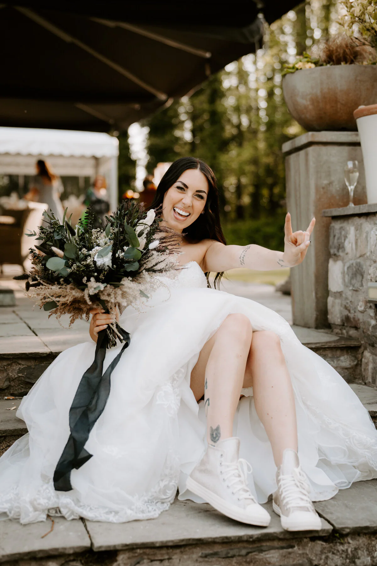 Bride in white Converse trainers flashing a rock-and-roll hand sign on stone steps with a dried bouquet.