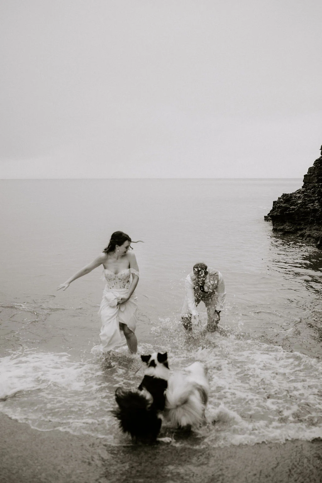 Black and white photograph of Alicia and Martin splashing through shallow water with their dogs