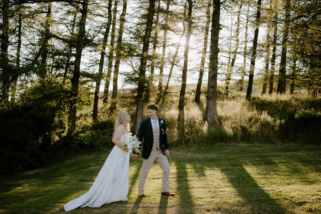 Ella and Josh standing together among tall trees in warm light