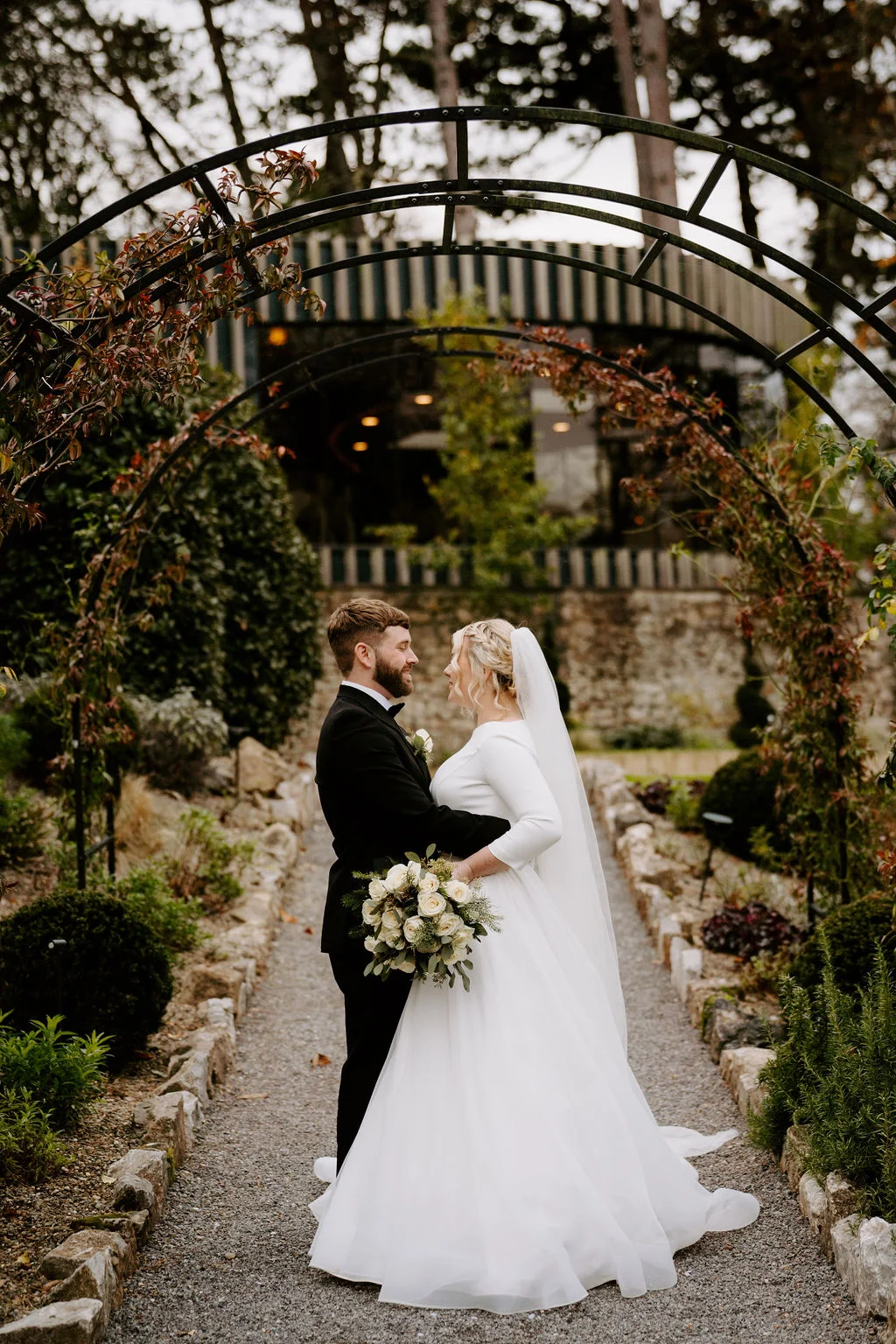 Max and Gwen embracing beneath a garden arch at Fairyhill