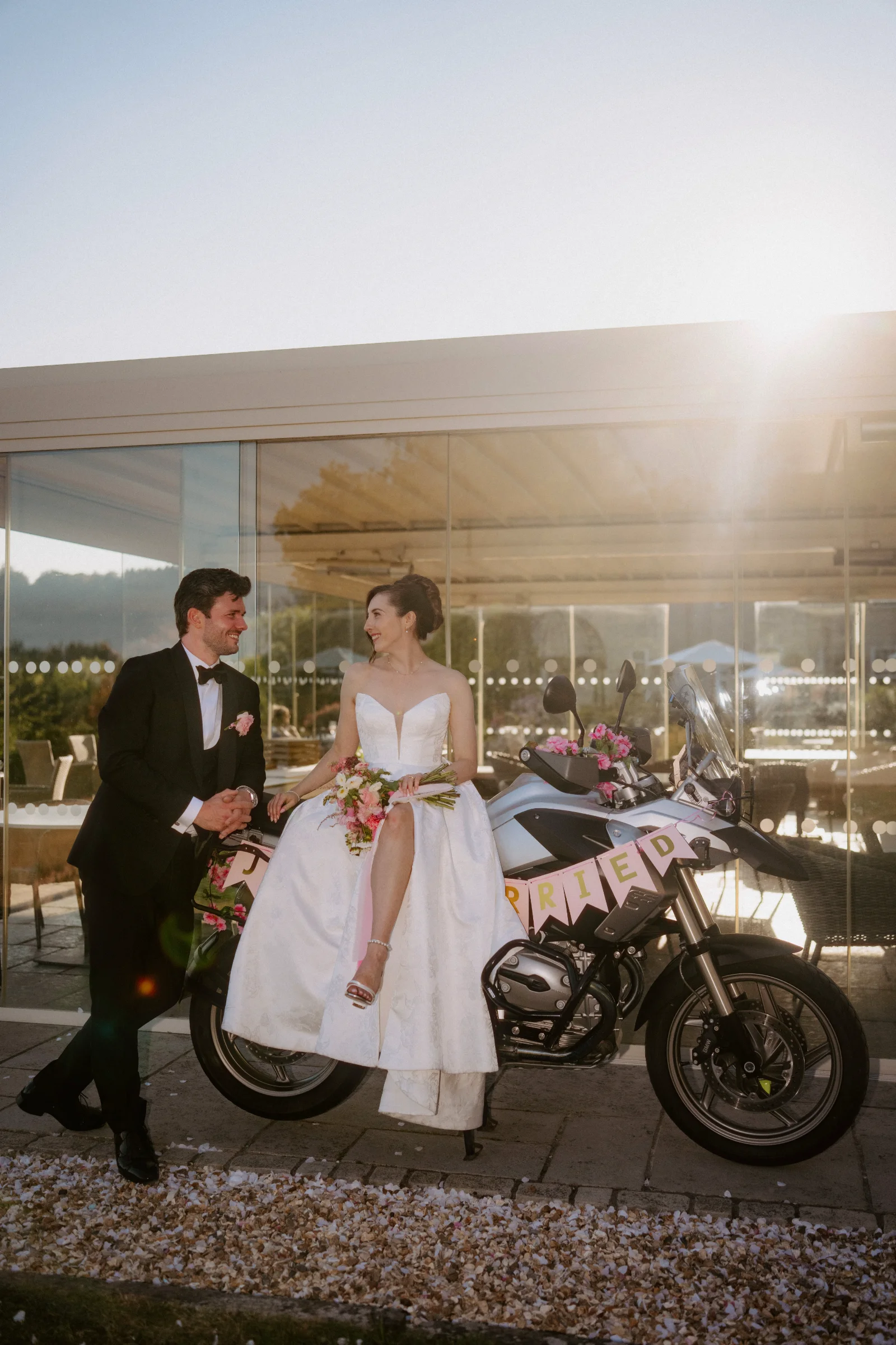 Newlyweds standing beside a vintage motorcycle outside their wedding venue.