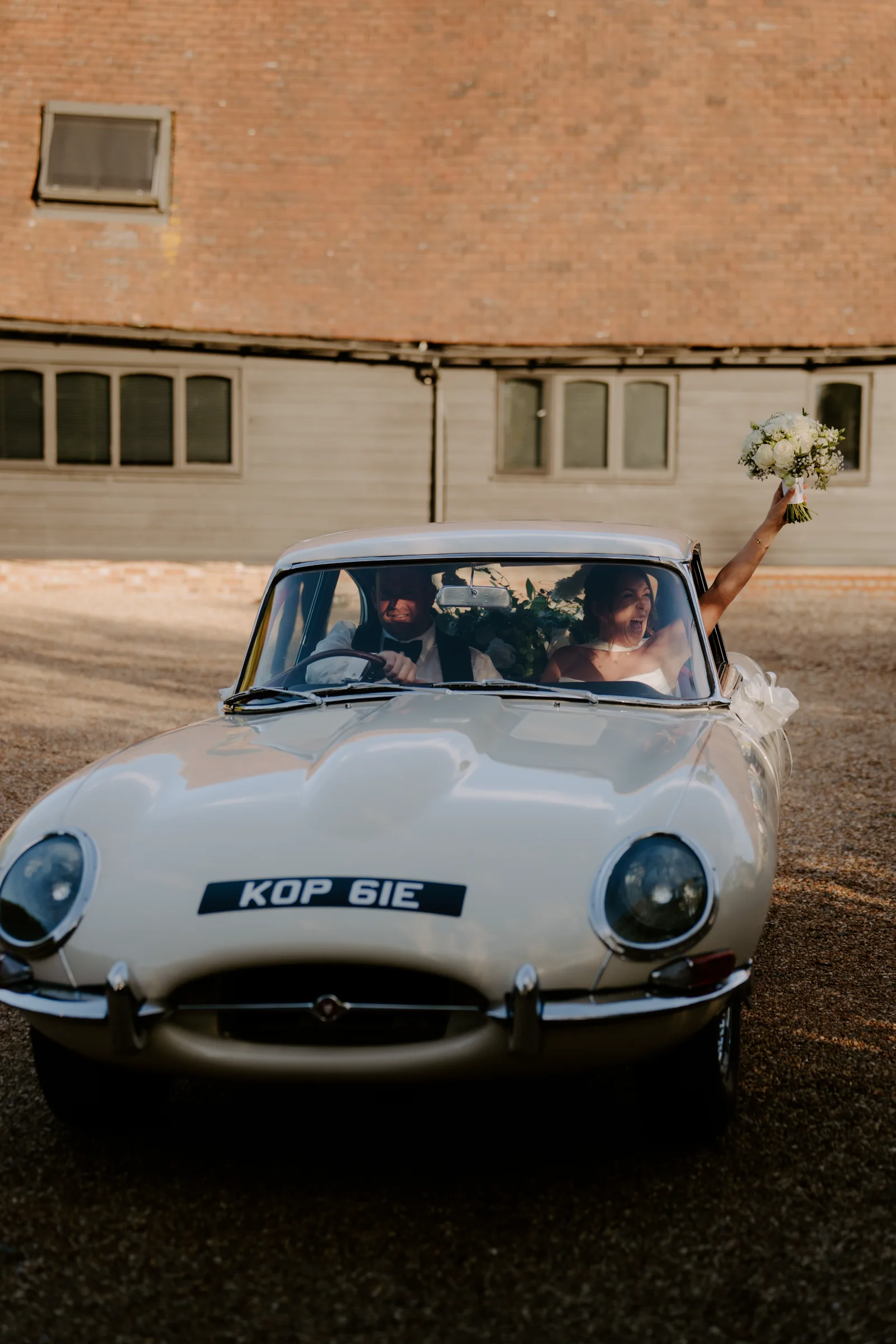 Newlyweds waving from a classic wedding car outside their venue.