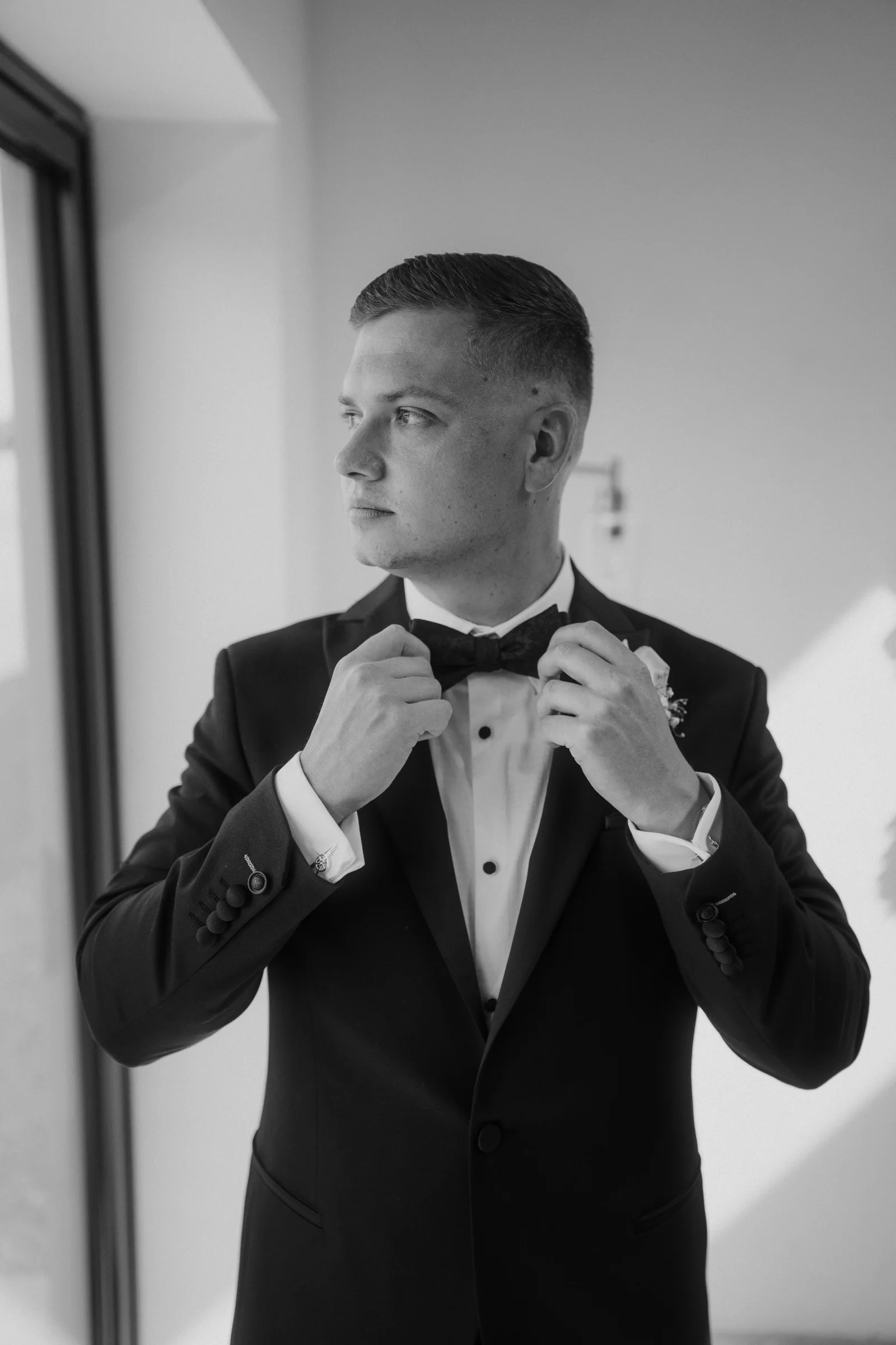 Black and white portrait of a groom adjusting his bow tie before the ceremony.