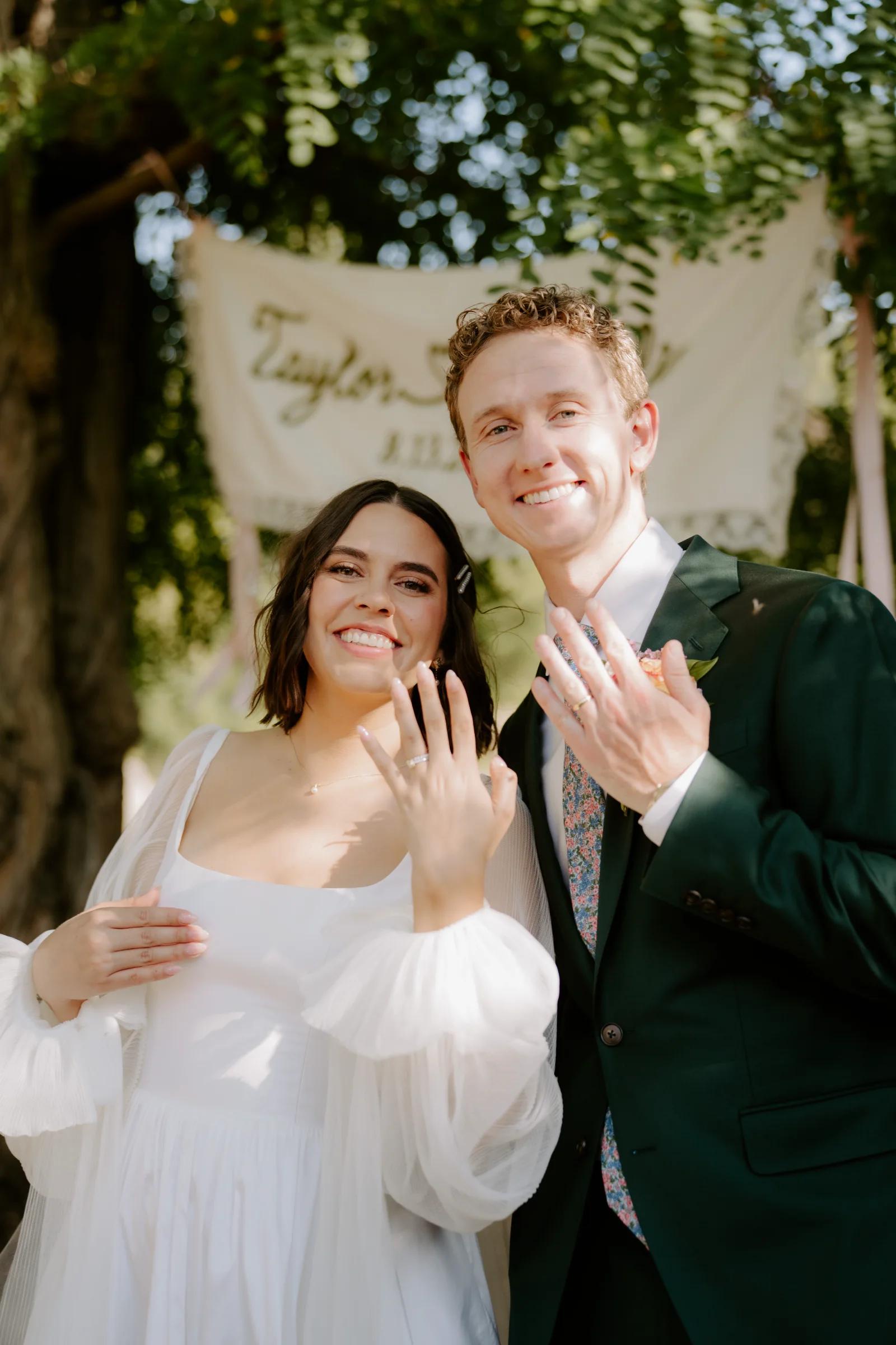 Newlywed couple smiling and showing their wedding rings beneath a floral arch.