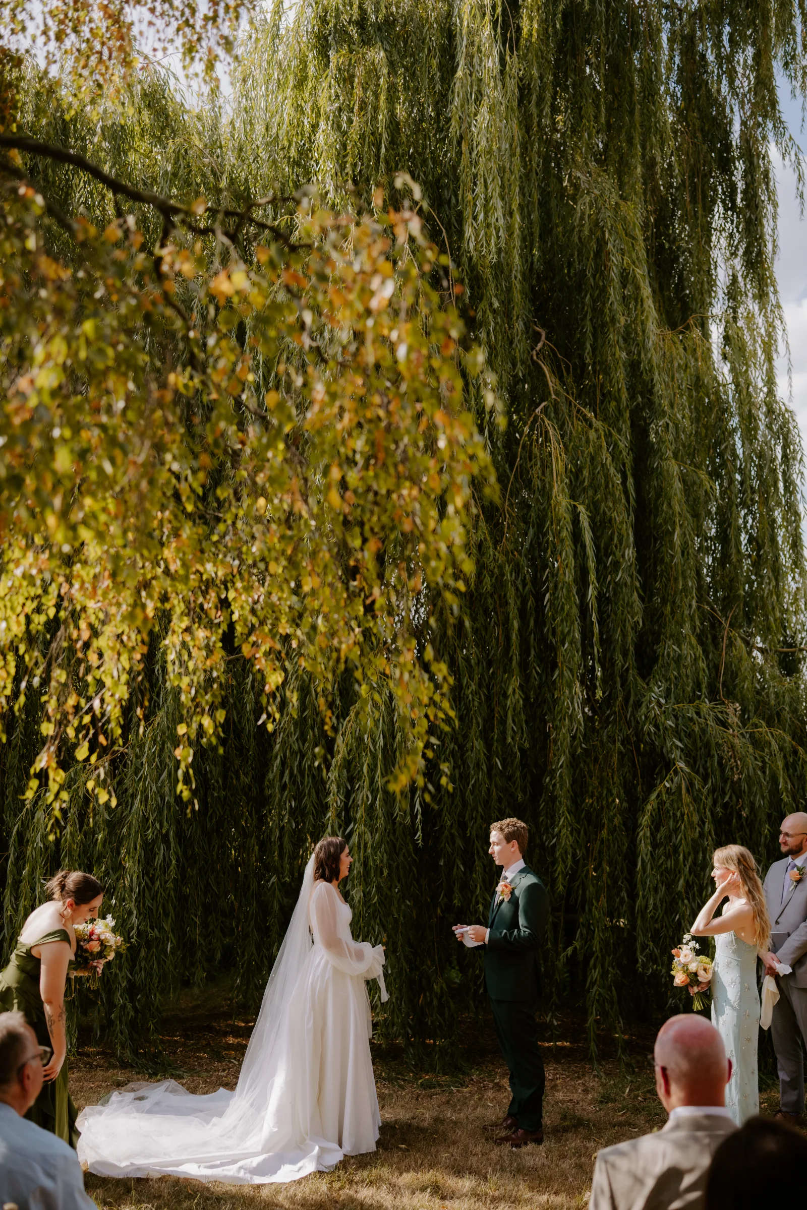 Outdoor wedding ceremony beneath a weeping willow tree with guests gathered around.