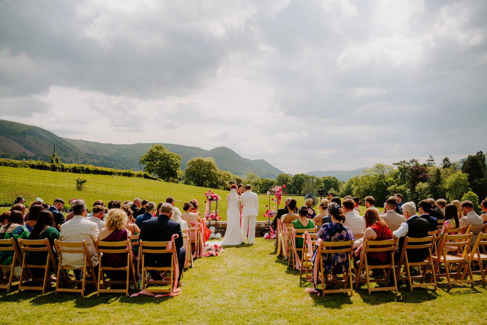 Molly and Isobel stand at the end of an aisle of wooden chairs on a lawn during their outdoor ceremony, with guests seated either side and green hills behind