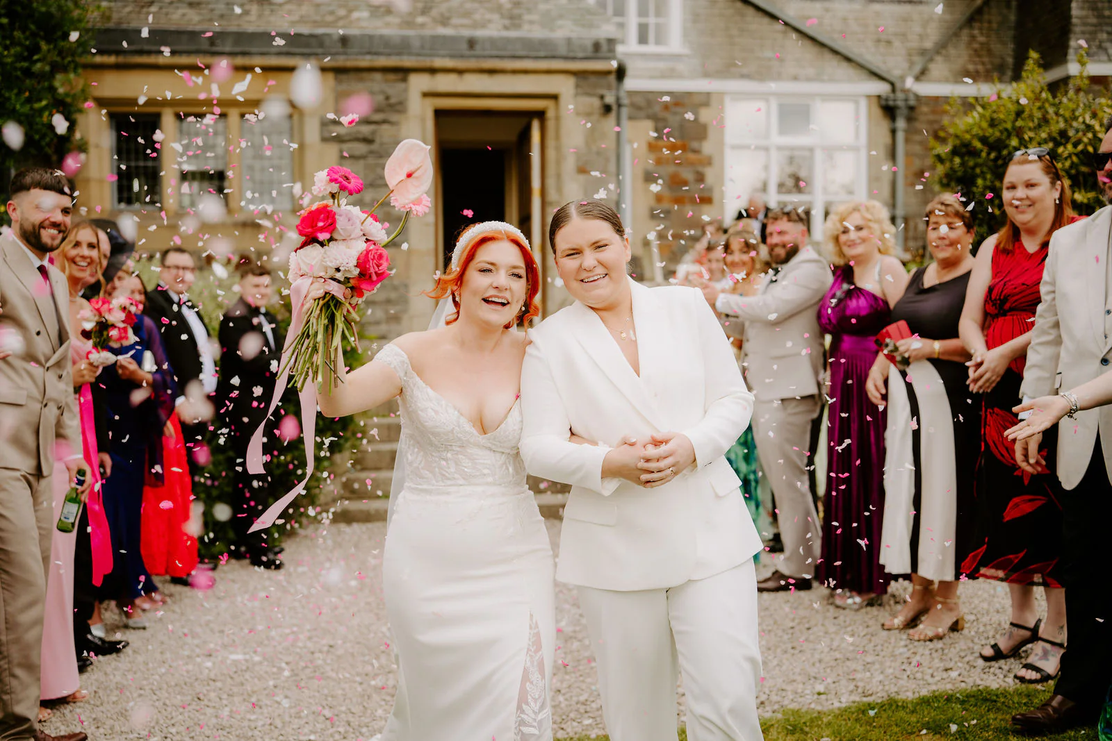 Molly and Isobel walk arm in arm through a shower of confetti thrown by guests lining a gravel path in front of the stone house at Plas Dinam, one in a lace gown and one in a white suit