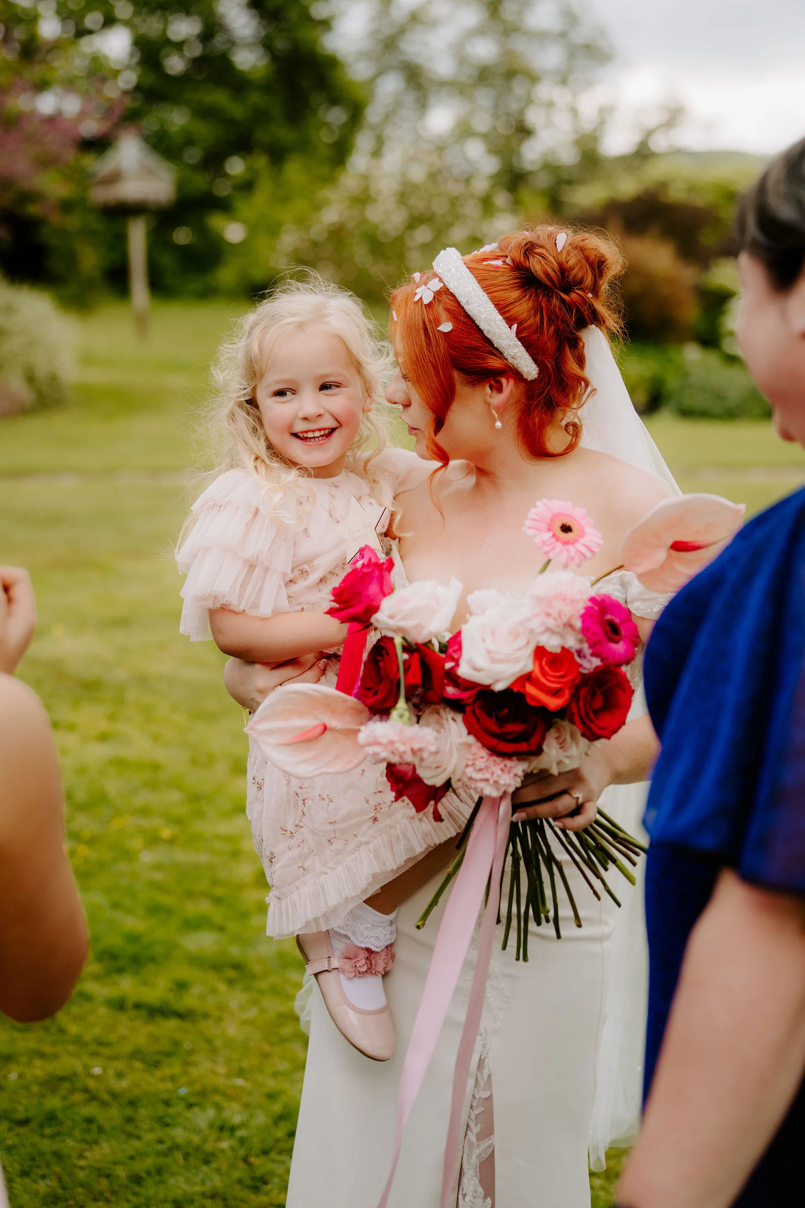 One of the brides, in a veil and headband, smiles as she carries a small child in a pale pink dress and holds a bouquet of pink and red flowers on the lawn