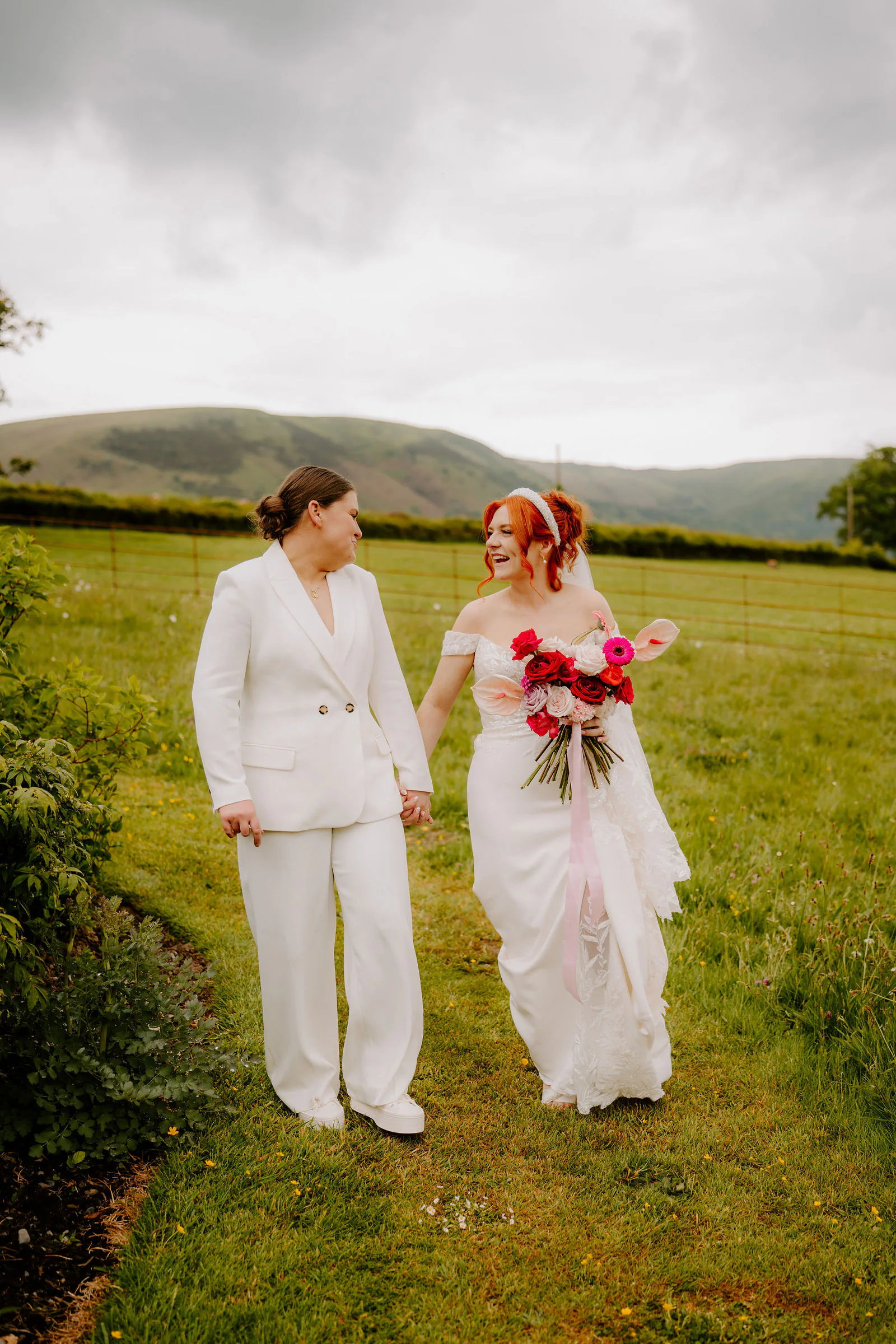 Molly and Isobel, one in a white suit and one in a lace gown with a red and pink bouquet, walk together along the grass with a ridge of hills behind them