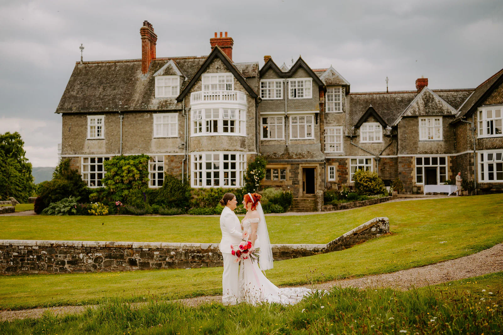 Molly and Isobel embrace on the lawn in front of the gabled stone house at Plas Dinam, with a low stone wall and a winding path in front of them