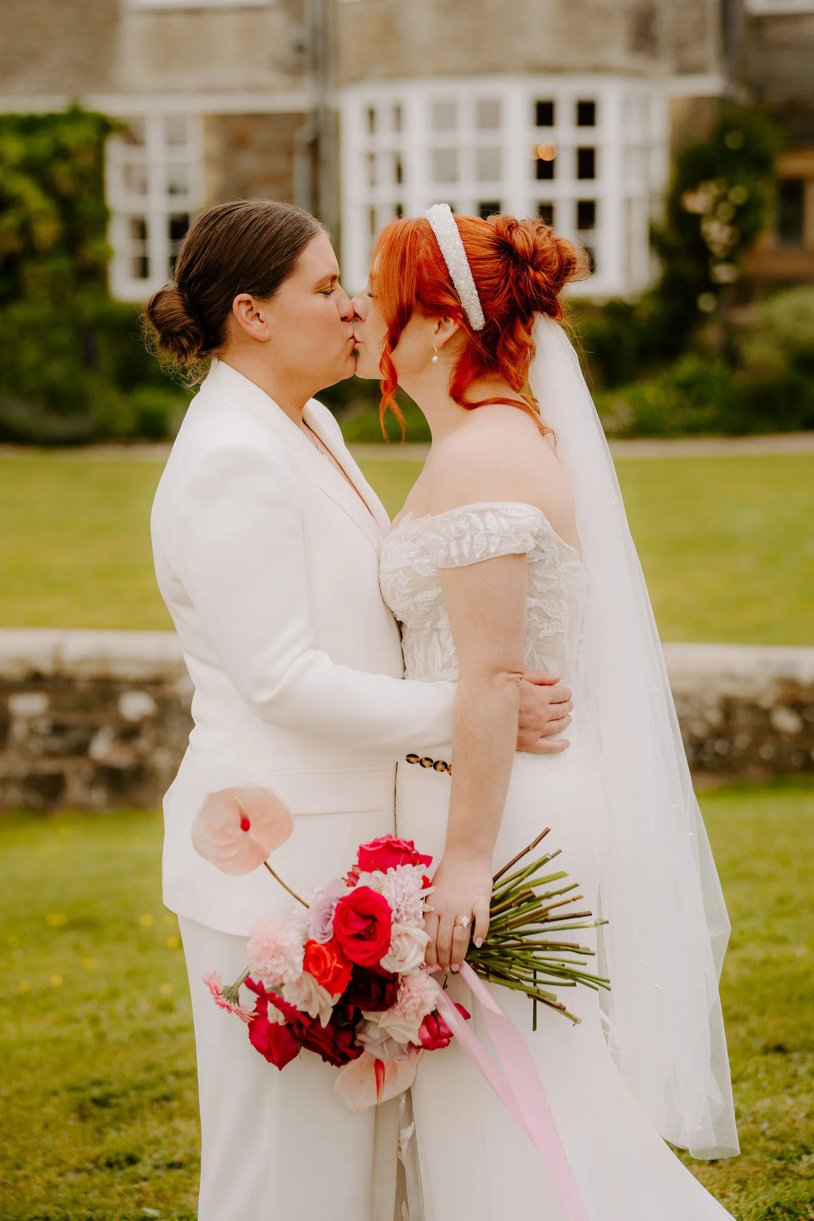 Molly and Isobel kiss on the lawn, one in a white suit and one in an off-the-shoulder gown and long veil, with a bouquet of anthuriums and roses between them