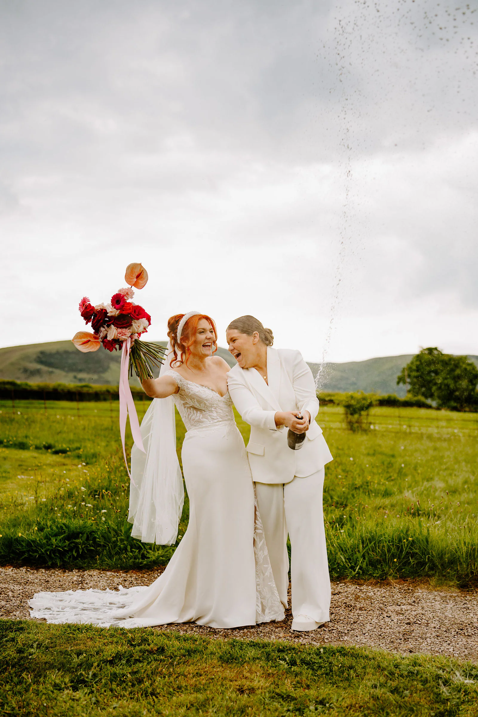 Molly and Isobel laugh as champagne sprays from a bottle on a gravel path, one raising a bouquet, with hills behind them