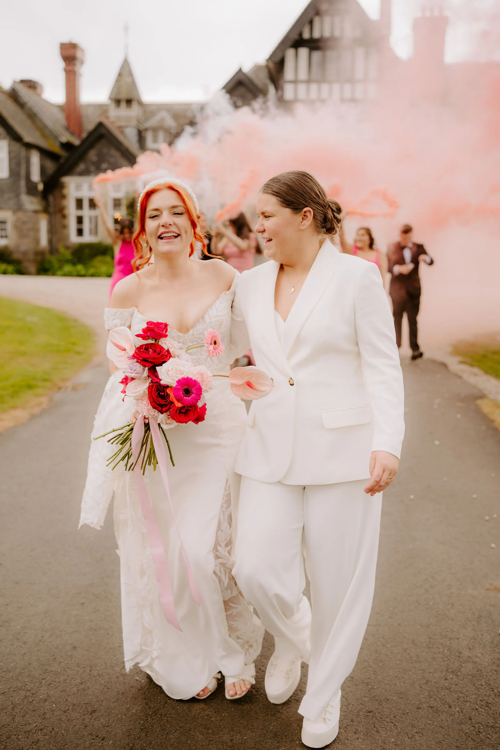 Molly and Isobel walk down a drive laughing with pink smoke billowing behind them in front of a half-timbered wing of the house