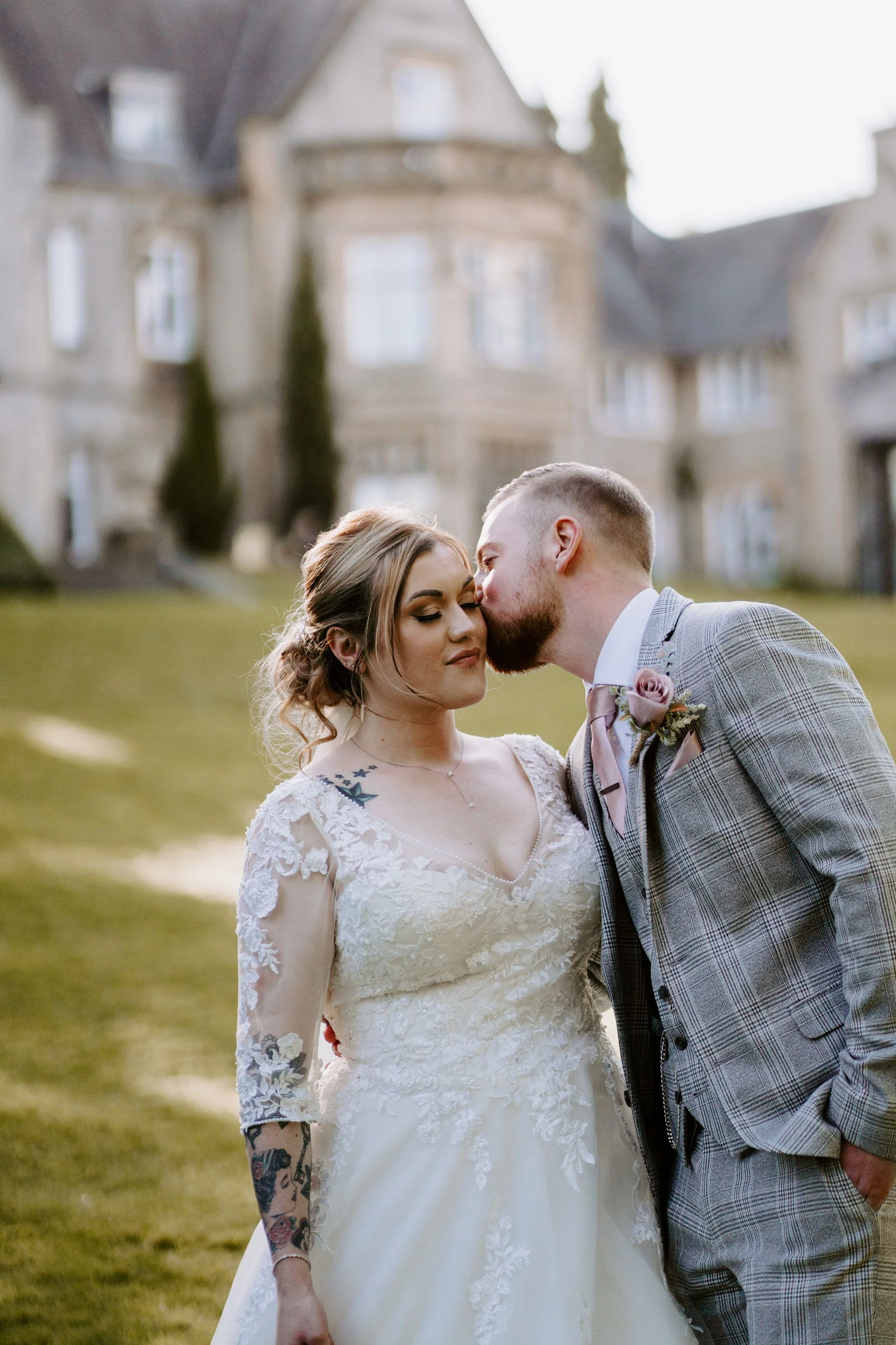 A groom in a grey check suit kisses his bride on the cheek, with a stone hall softly out of focus behind them