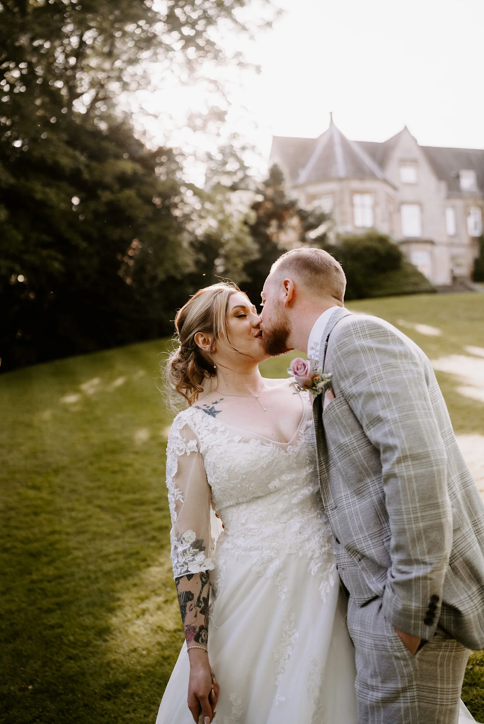 A bride in a lace-sleeved gown and a groom in a grey check suit kiss on a lawn with the pointed roofs of a stone hall behind