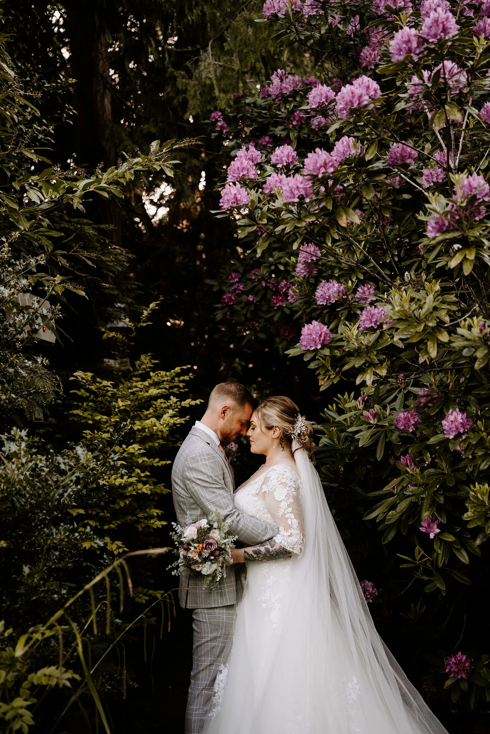 A bride in a long veil and a groom in a grey check suit stand forehead to forehead against a bank of purple rhododendron